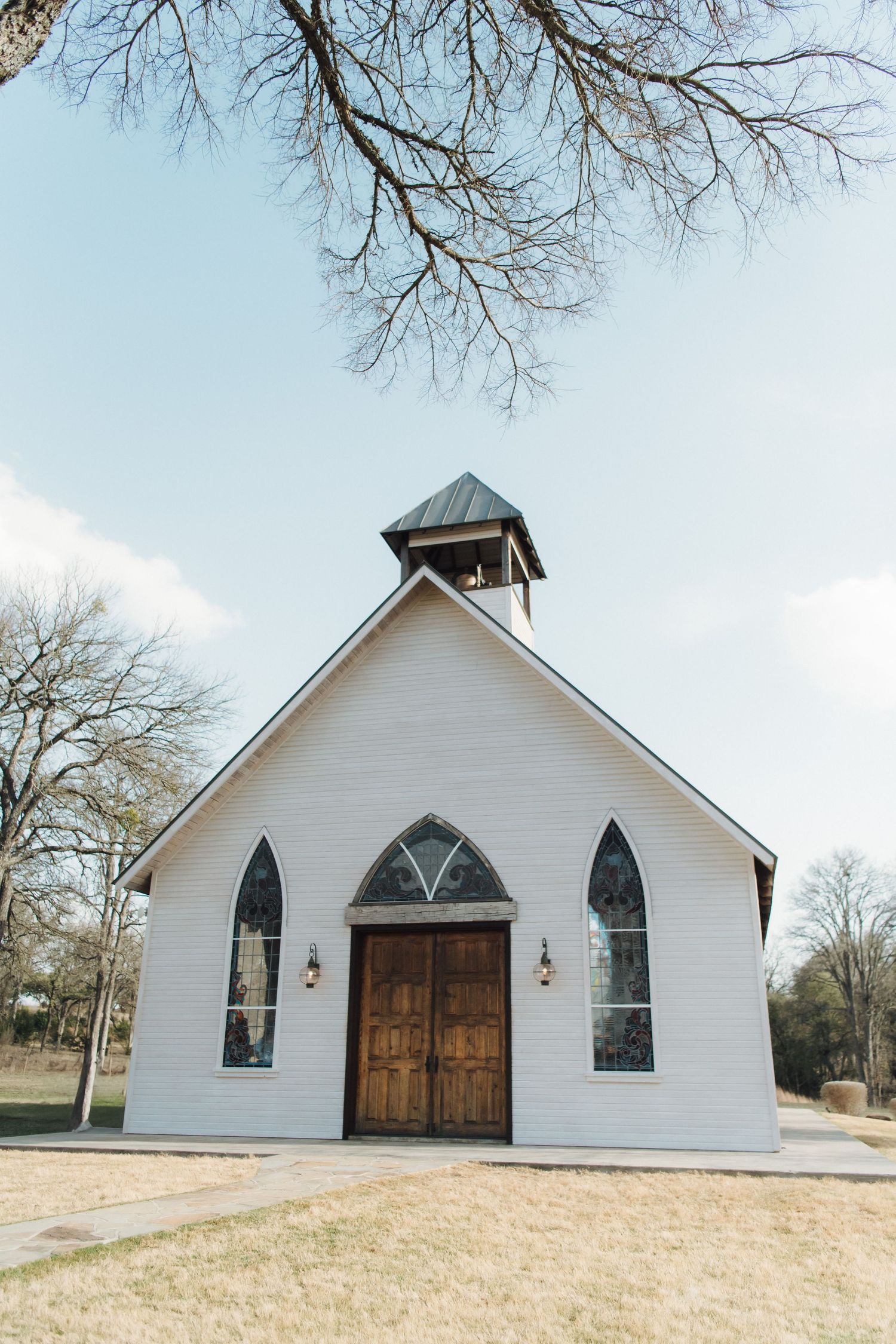A charming white country chapel with arched windows, wooden doors and a small bell tower stands against a light blue sky.