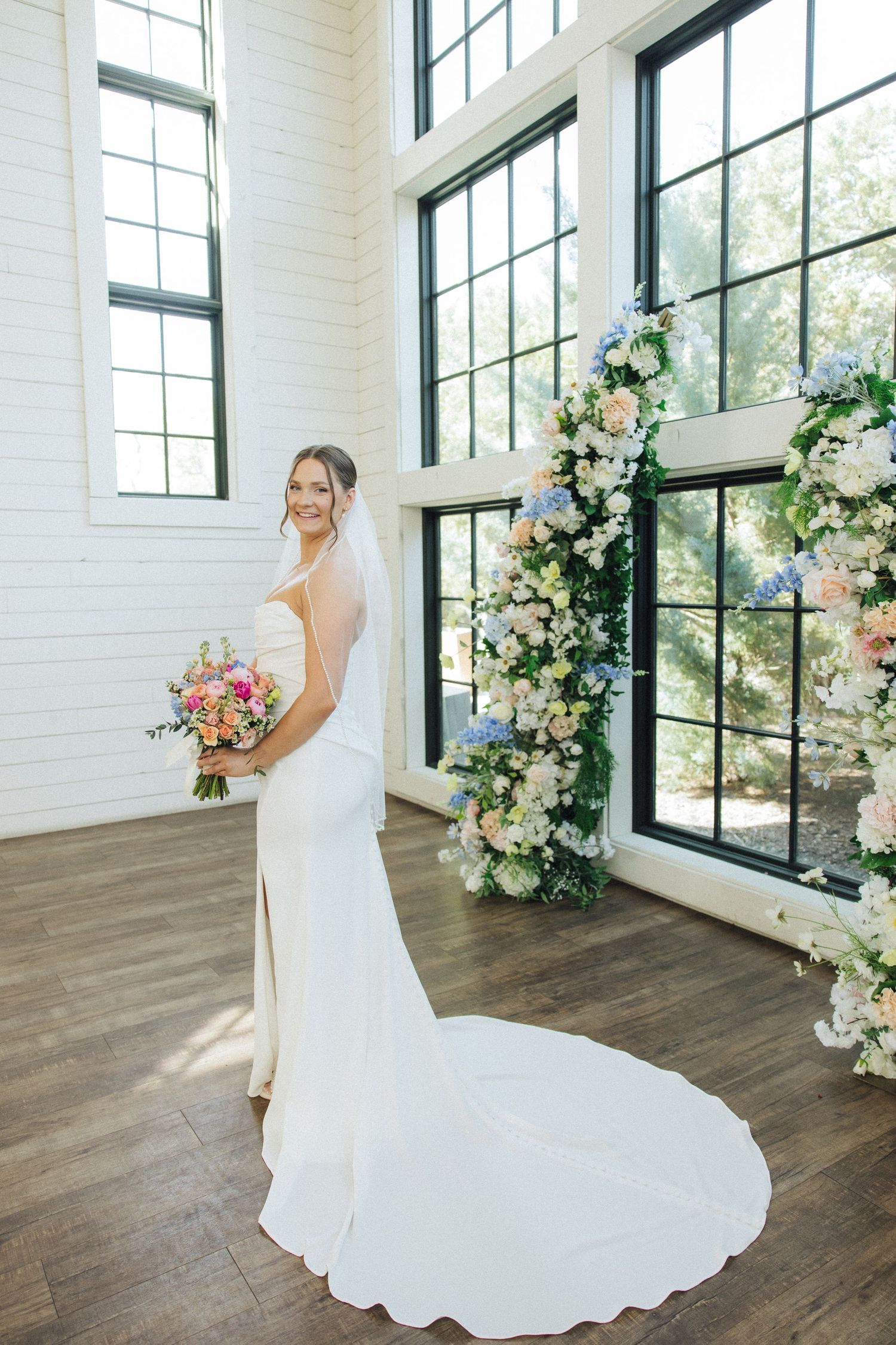 Beautiful wedding dress with flowing train photographed in bright venue with floral decor and large windows.