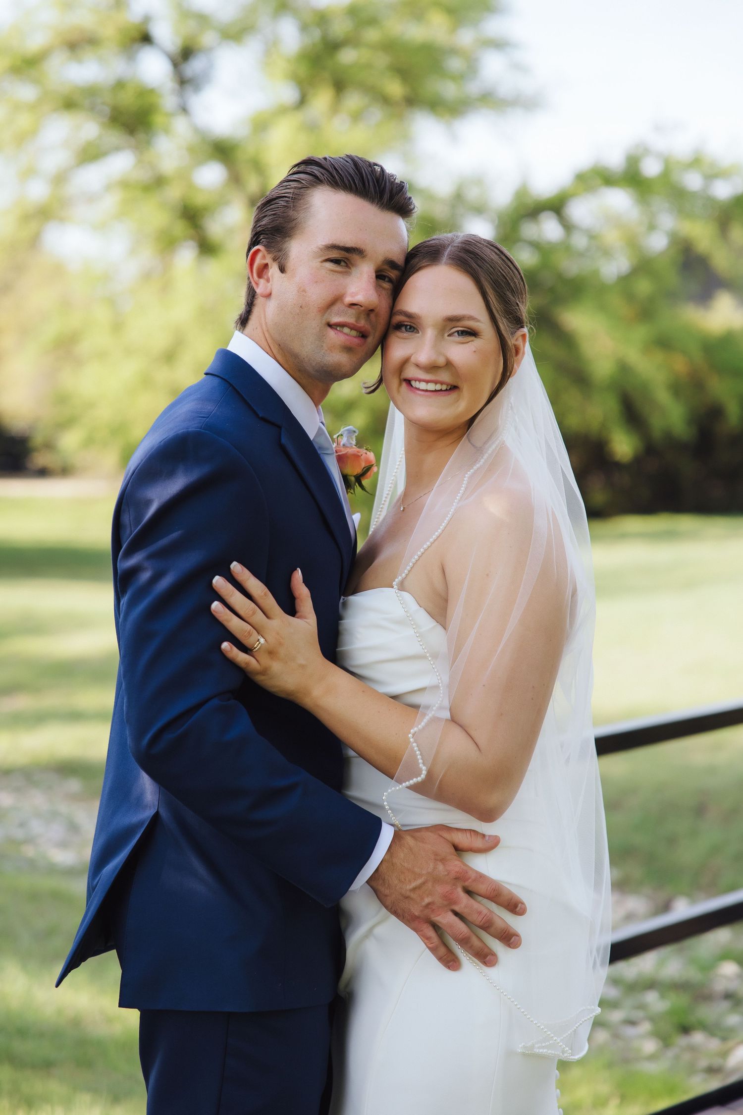 A bride and groom embrace outdoors on their wedding day, she in white and he in navy, surrounded by greenery.