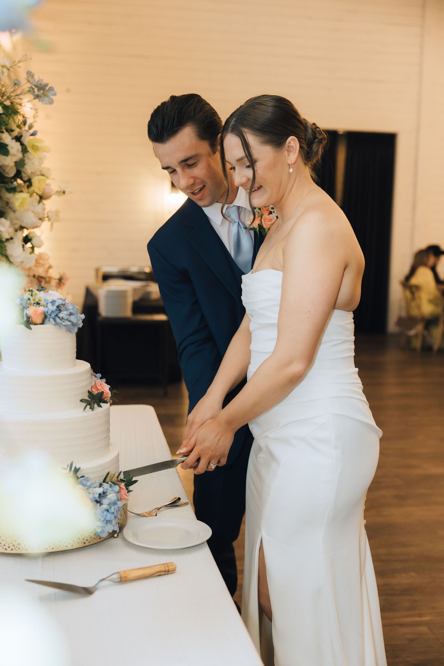 A couple cuts their wedding cake together at their reception in an indoor venue with white walls.