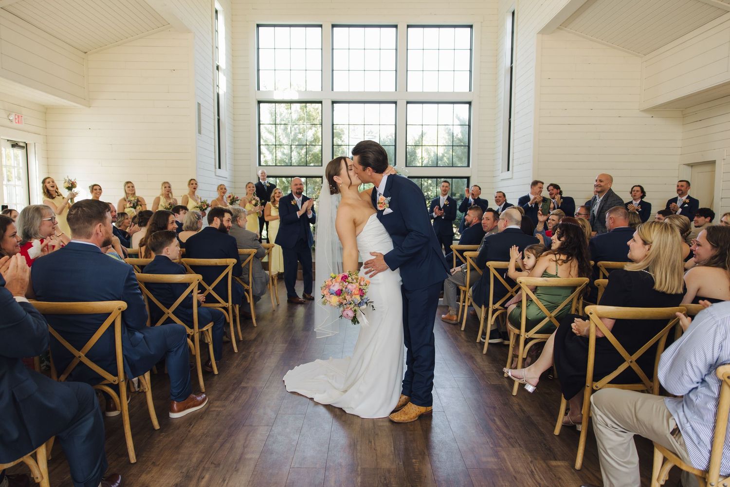 A couple shares their first kiss as newlyweds in a bright, airy venue filled with guests seated in wooden crossback chairs.