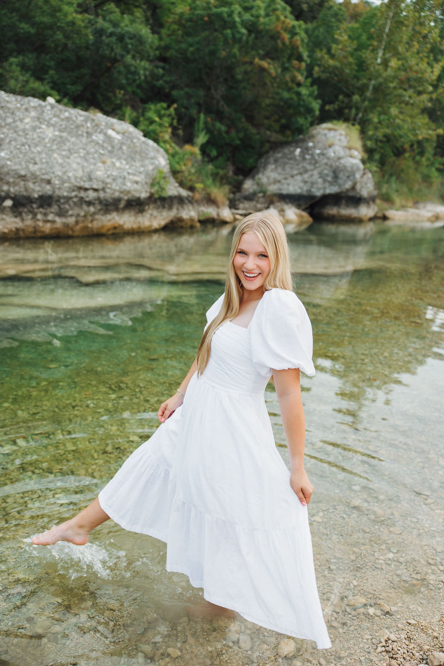 Someone in a white dress stands by crystal clear turquoise water near large limestone rocks in a natural setting.