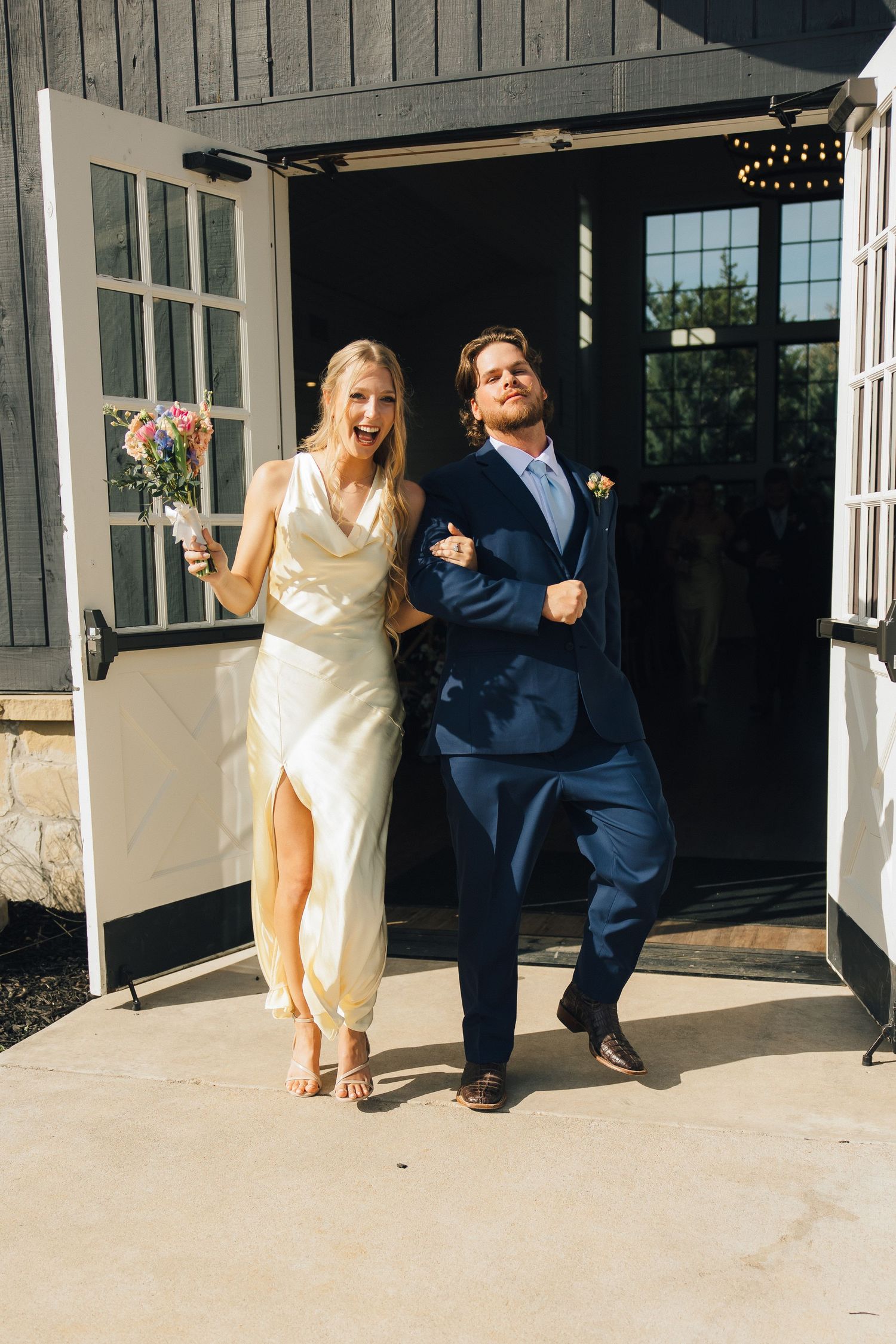 A couple in wedding attire joyfully exits their ceremony through a white-columned venue porch.