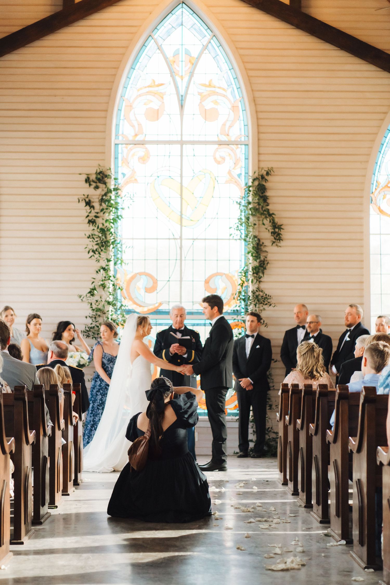 Wedding ceremony inside chapel with gothic arched window and greenery decorations along wooden church pews.