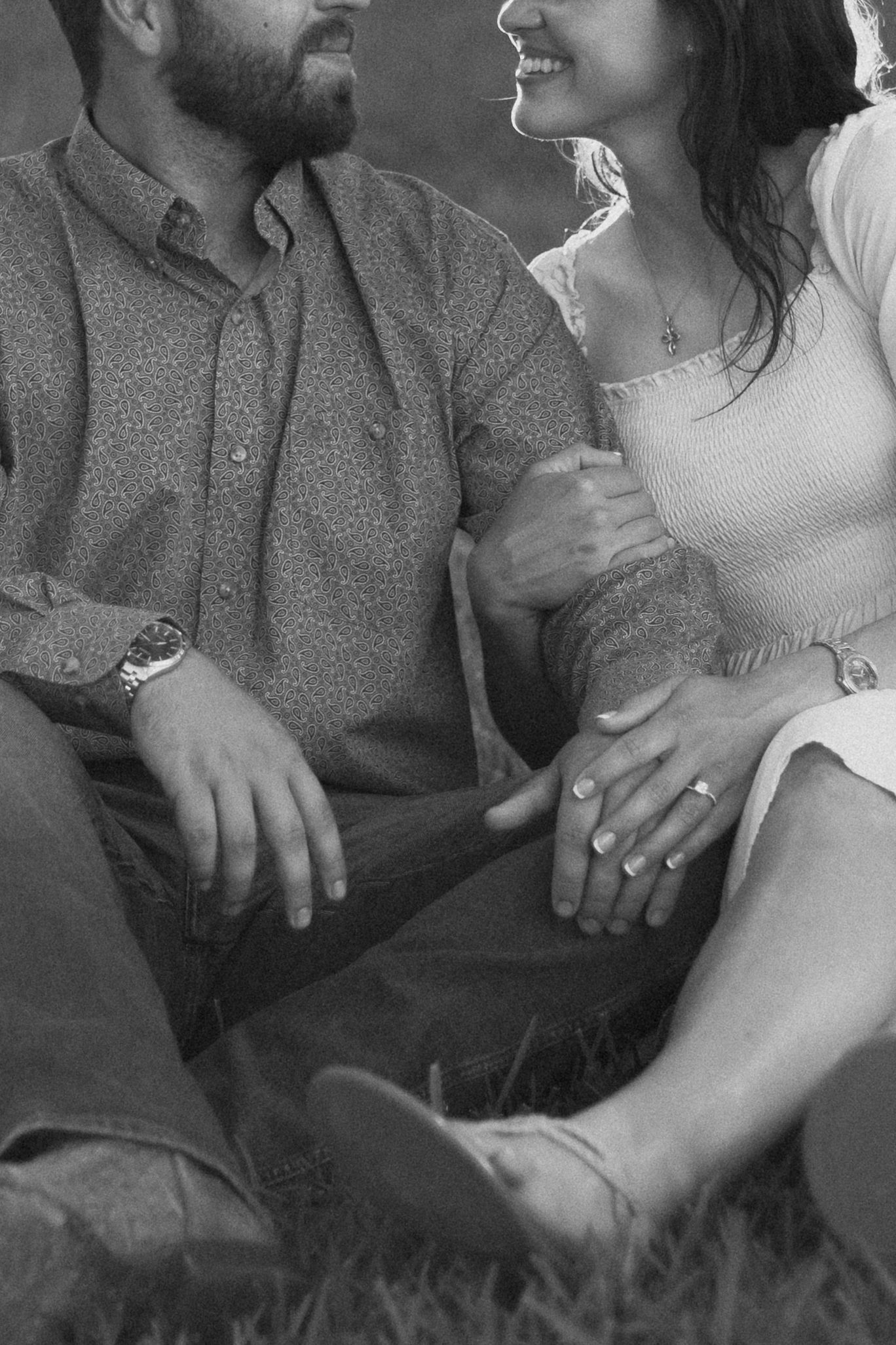 Black and white intimate portrait showing a couple sitting close together in casual clothing holding hands on grass.