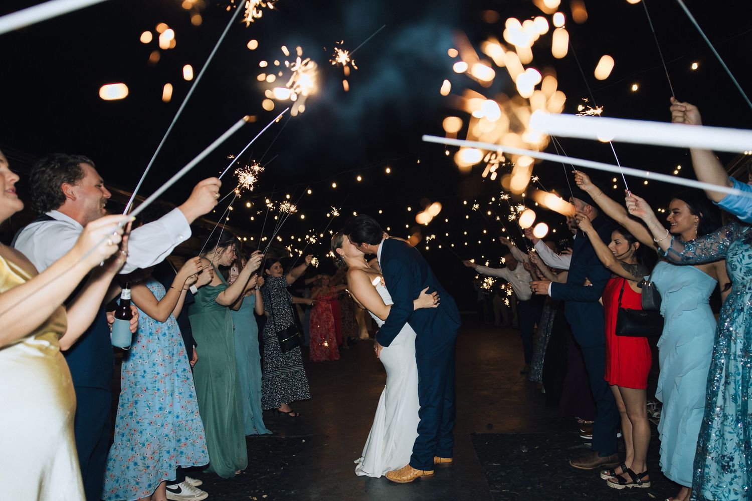 Wedding guests hold sparklers creating a magical nighttime tunnel effect during an outdoor celebration.