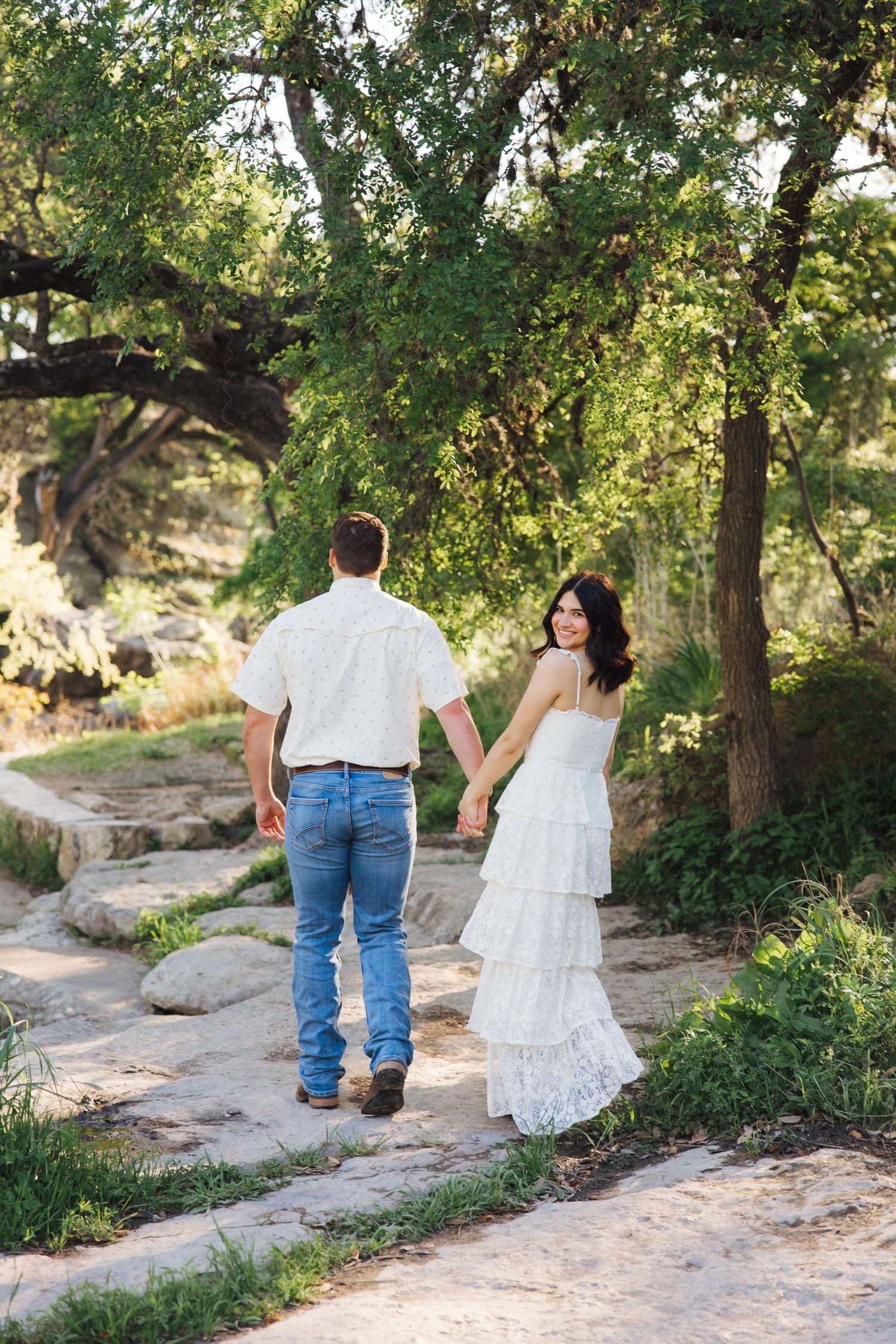 Two people walk hand in hand along a rocky path surrounded by lush green trees in a tranquil outdoor setting.