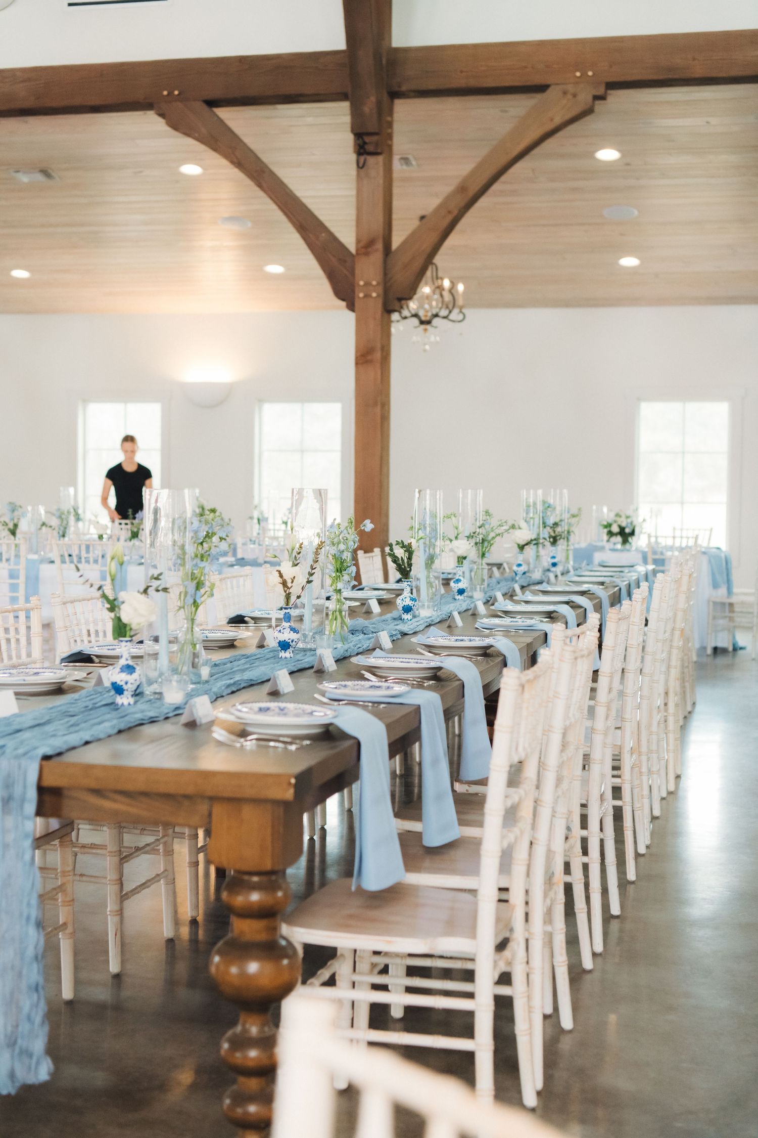 An elegant white wedding reception venue with exposed wooden beams features long tables decorated in soft blue and white theme.