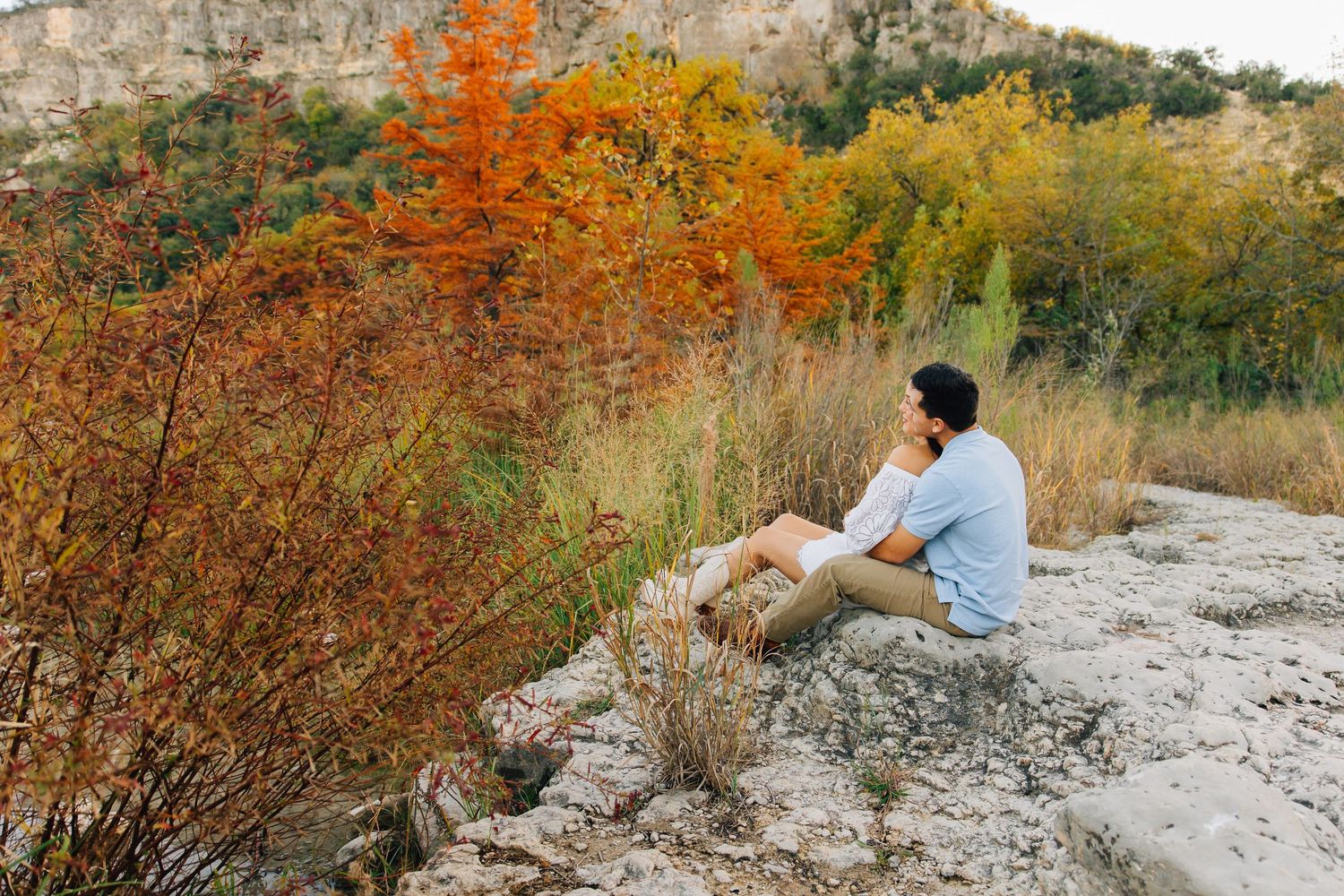 A person sits on rocky terrain surrounded by vibrant autumn foliage and golden trees against a mountainous backdrop.