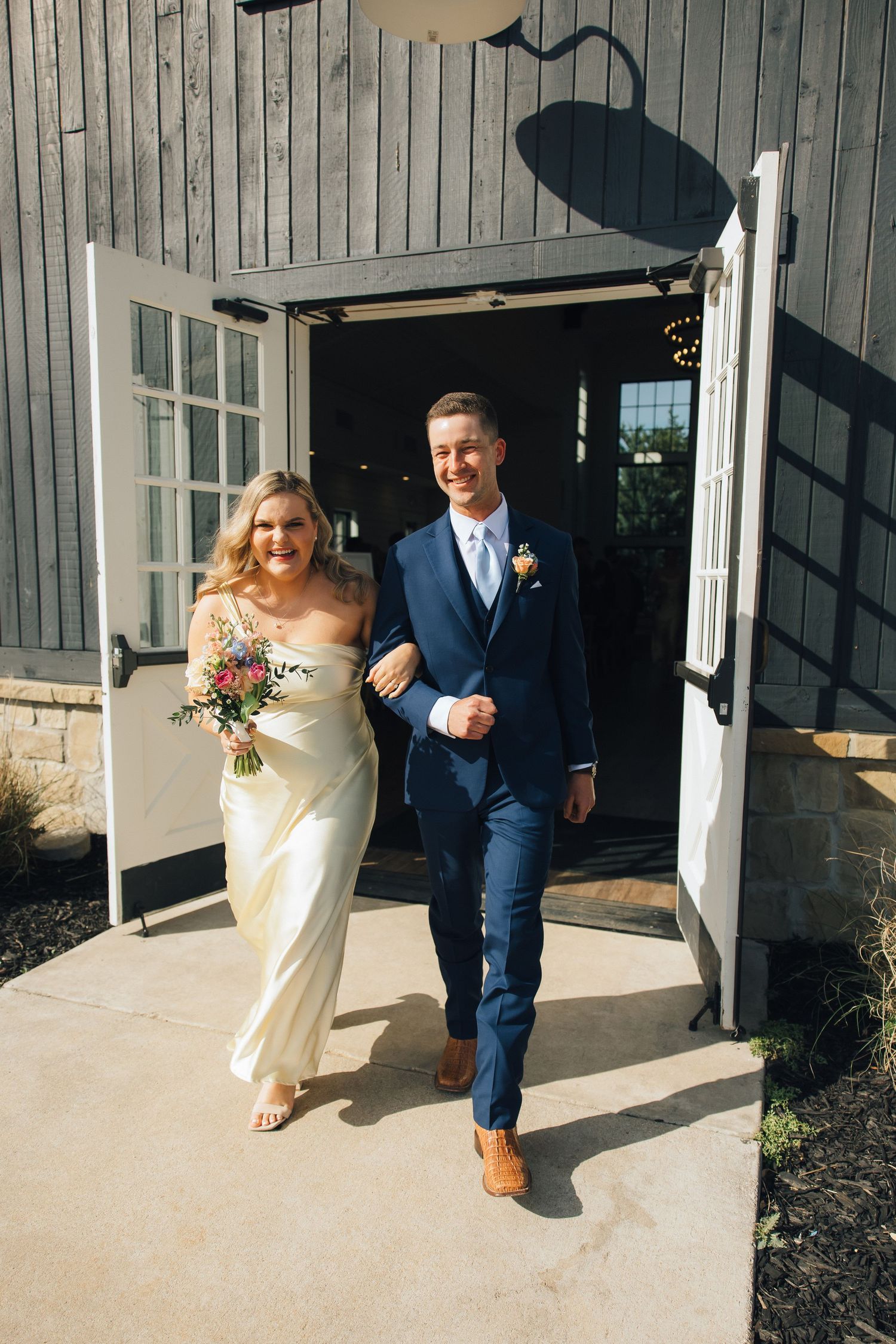 A wedding couple exits through white french doors of a gray barn venue with the bride in an off-shoulder dress.