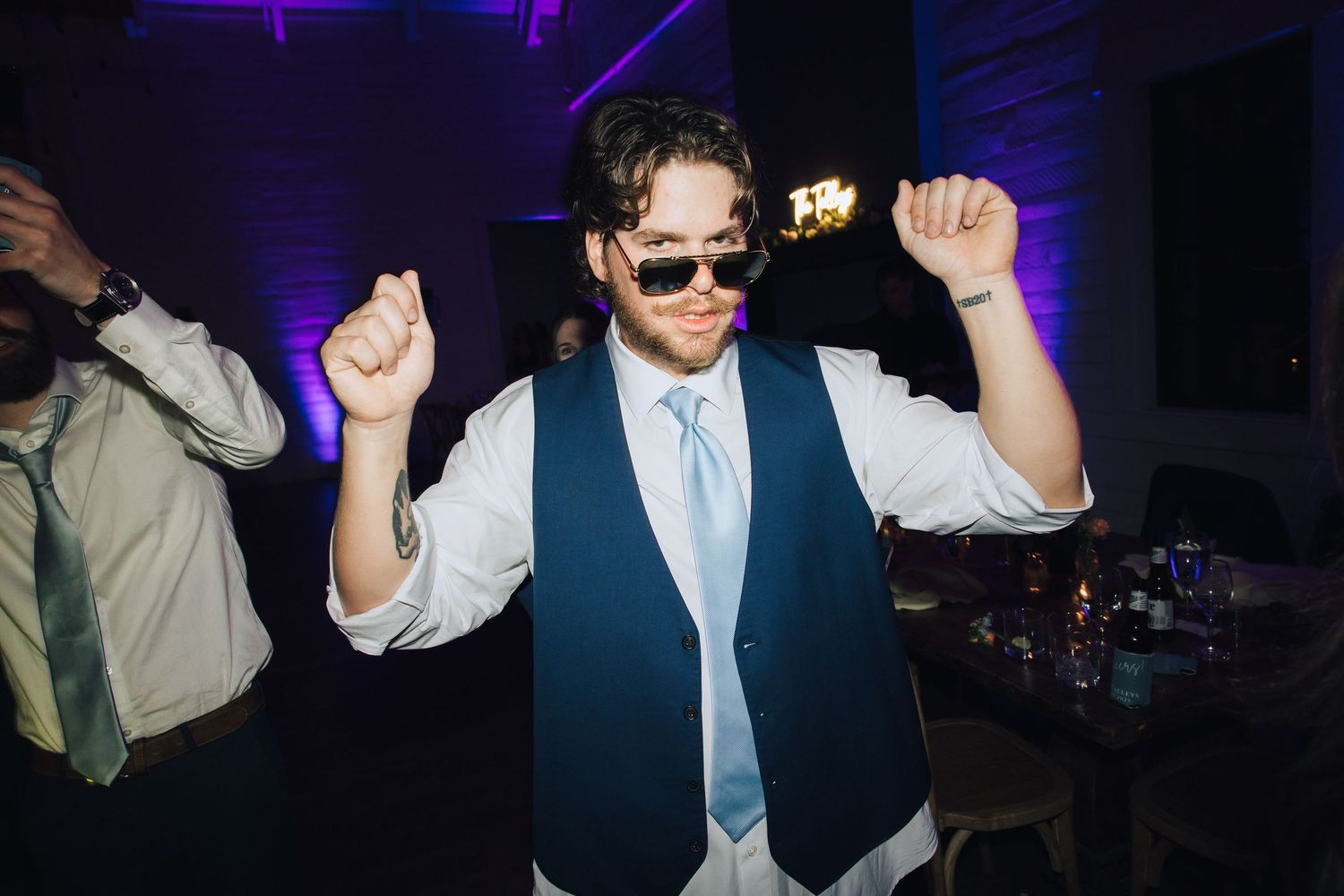 Person in a blue tie and vest dancing enthusiastically under purple event lighting.