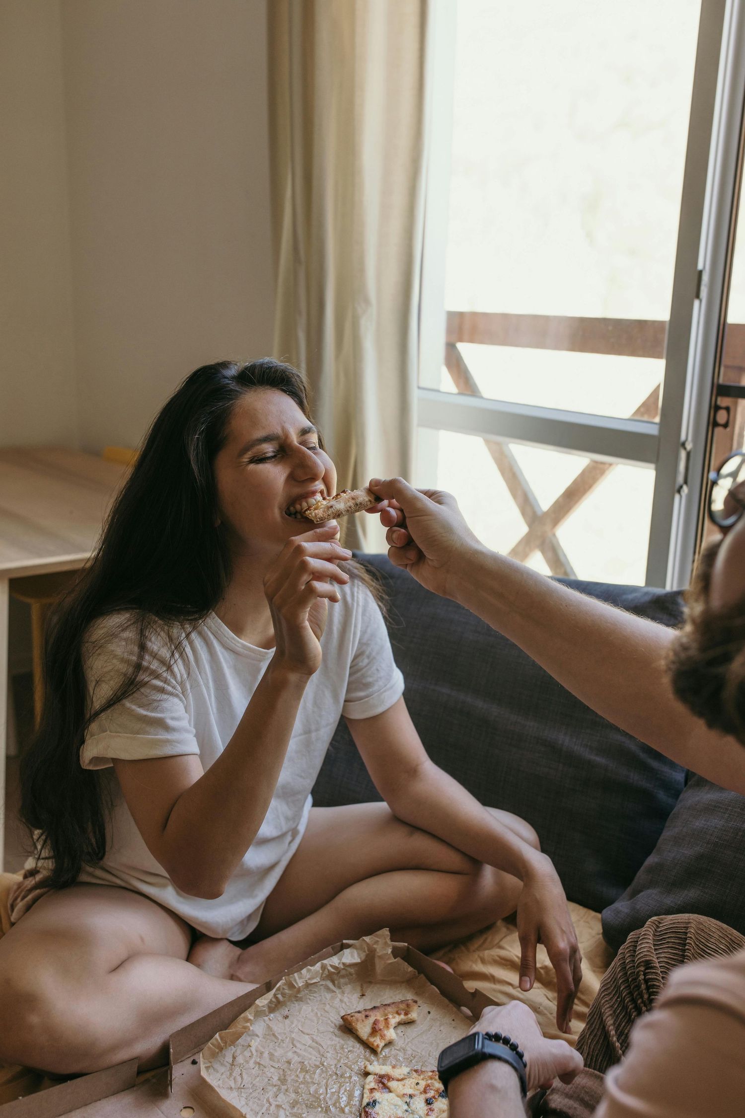 Two people sharing a casual meal together while sitting on the floor by a window in natural lighting.