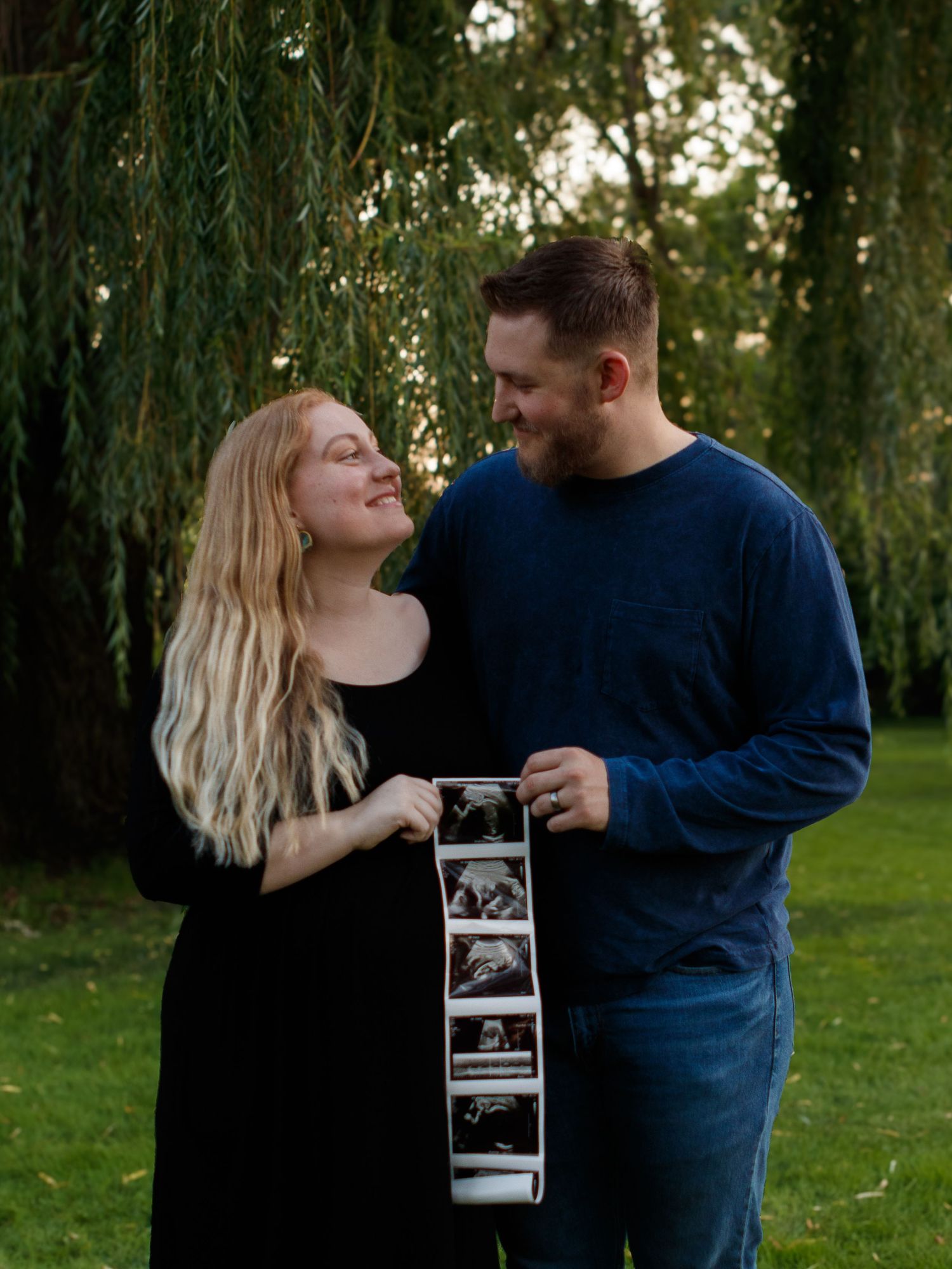 A couple shares an intimate moment while holding ultrasound photos under a willow tree in a park setting.
