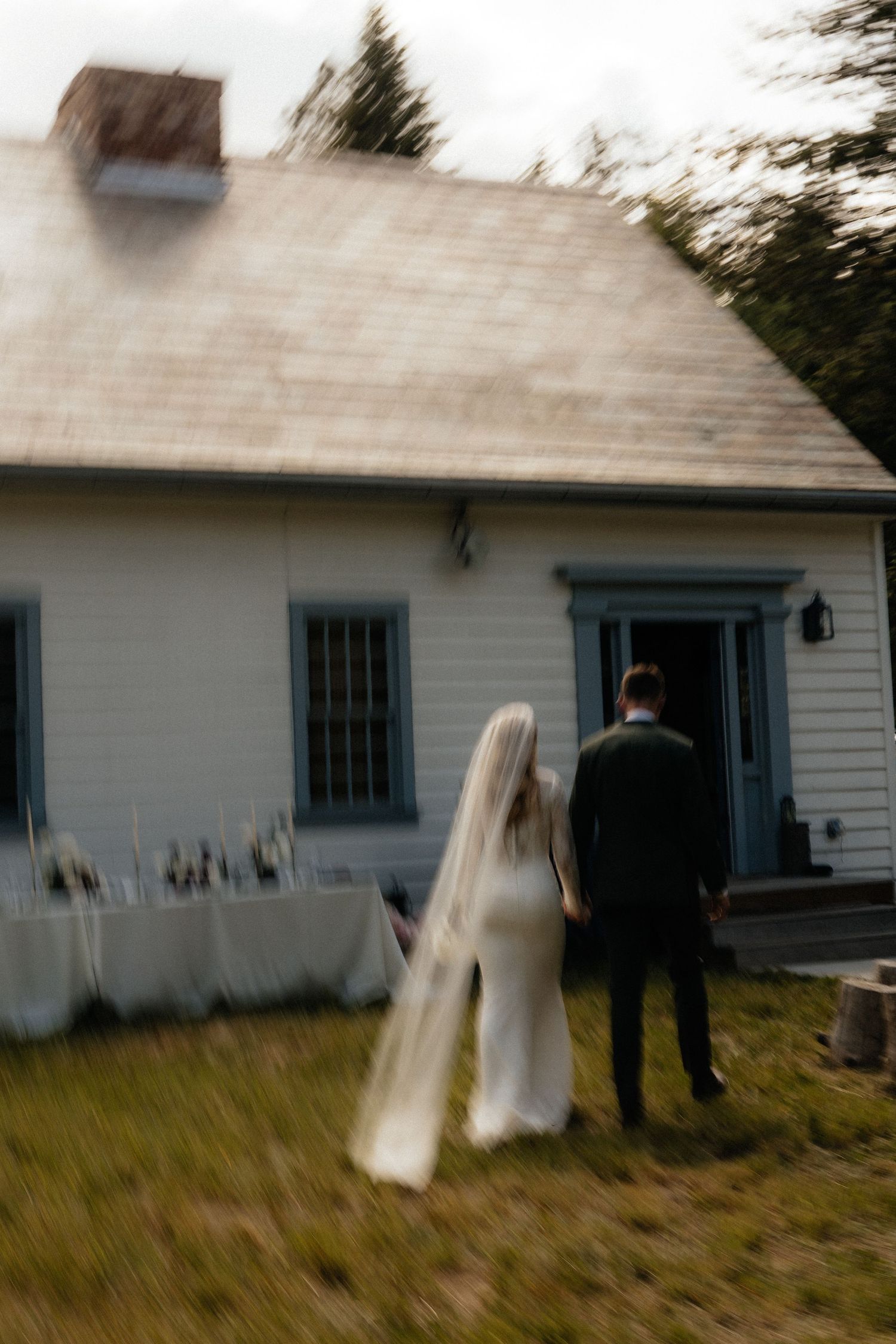 Couple in wedding attire stands in front of rustic white house with blue trim on grassy lawn in soft focus style.