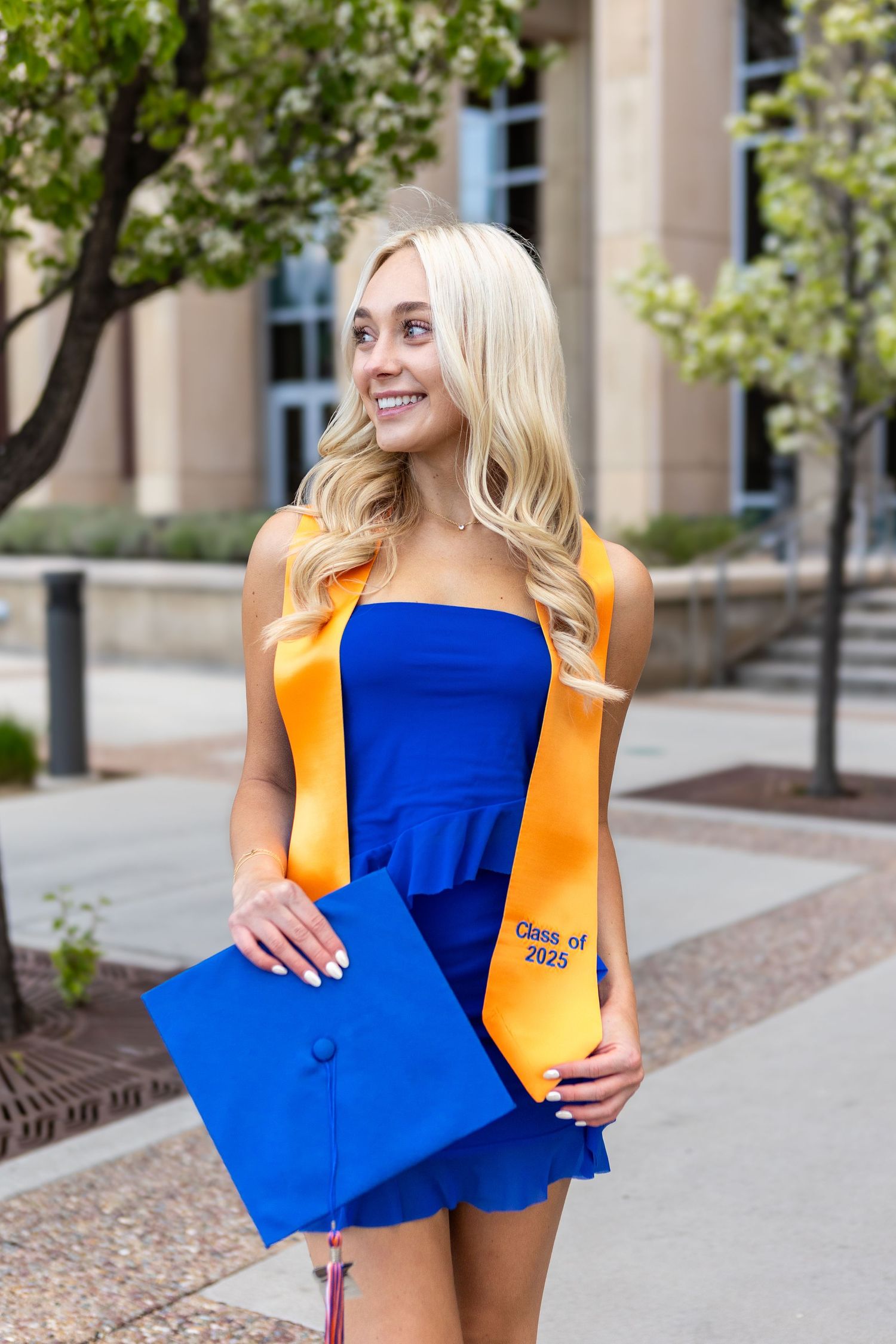Graduate in blue dress poses with bright orange stole and blue graduation cap on sunny campus grounds.