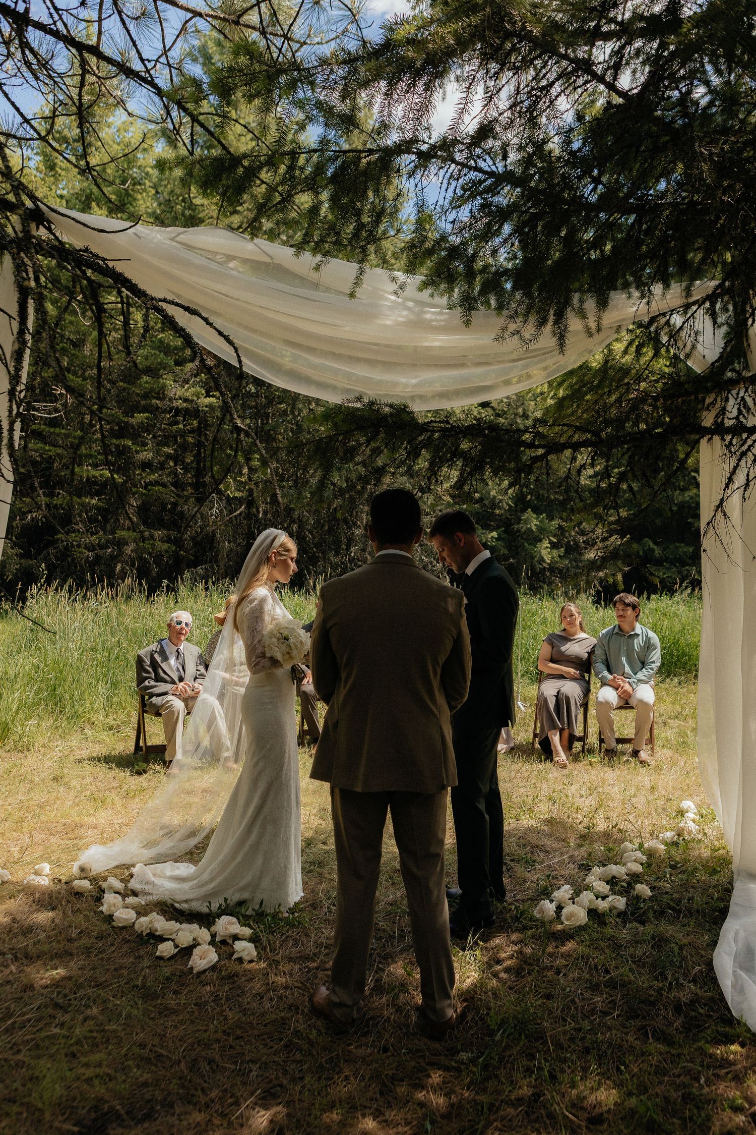 An intimate outdoor micro-wedding ceremony takes place under a draped canopy in a secluded forest setting.