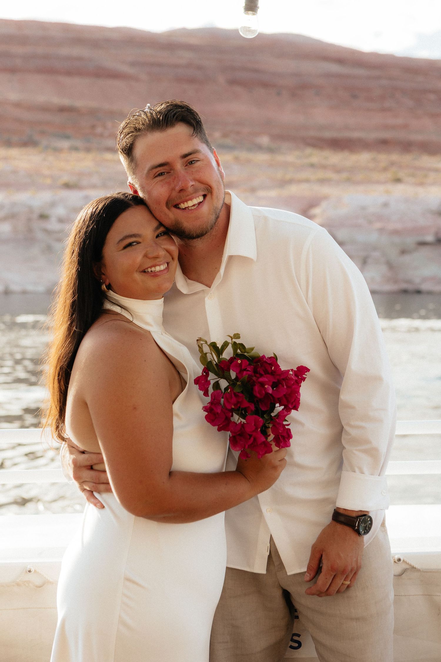 A couple stands in a desert landscape with pink flowers while sharing a romantic moment at sunset.