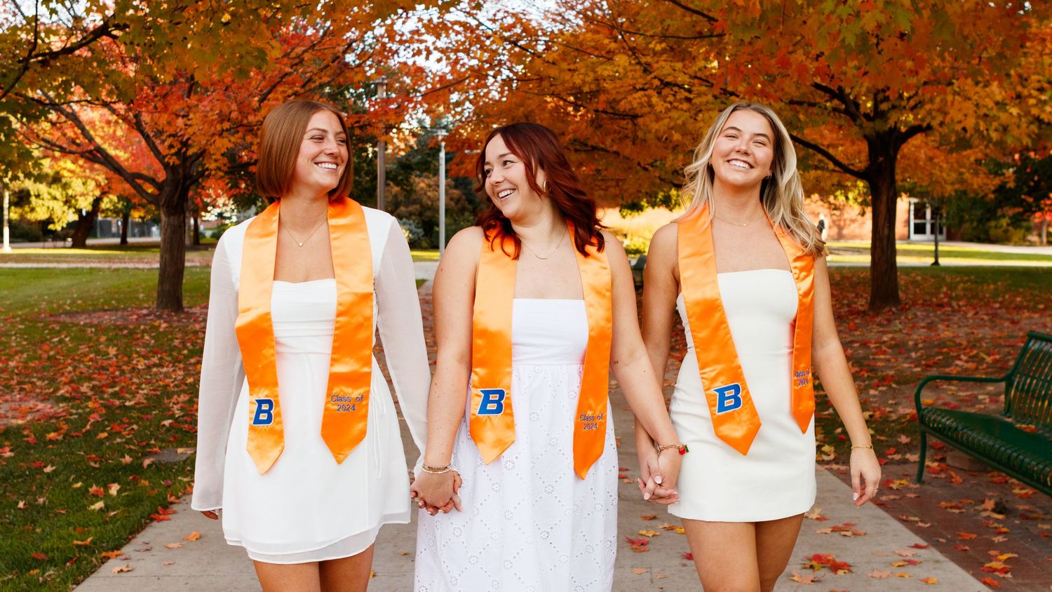 Three people wearing matching white dresses with orange sashes walk together on a path surrounded by autumn trees.