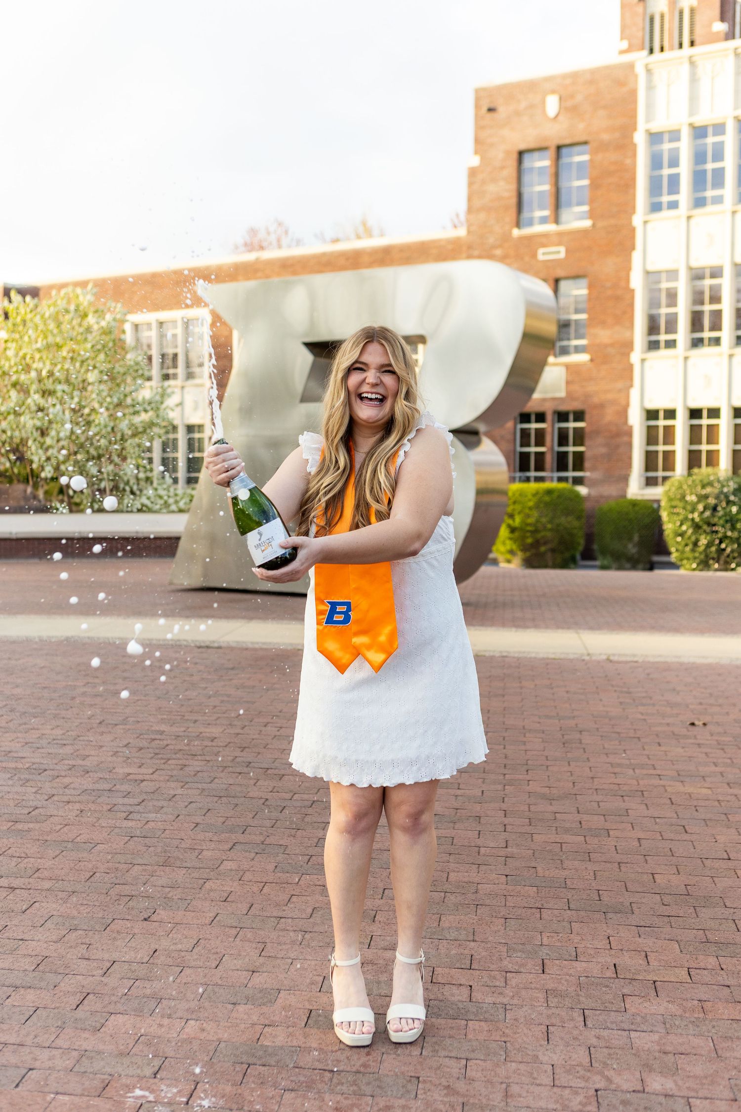 Graduate celebrating on campus brick plaza with champagne bottle and orange sash in white lace dress and sandals.