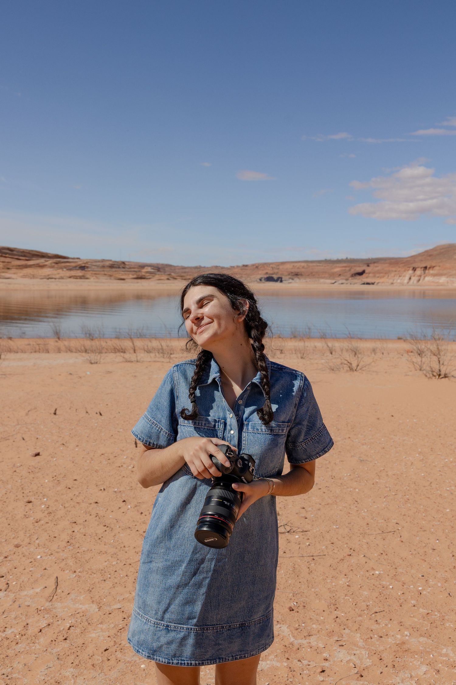 Photographer stands by scenic desert lake holding camera wearing denim dress in warm afternoon light.
