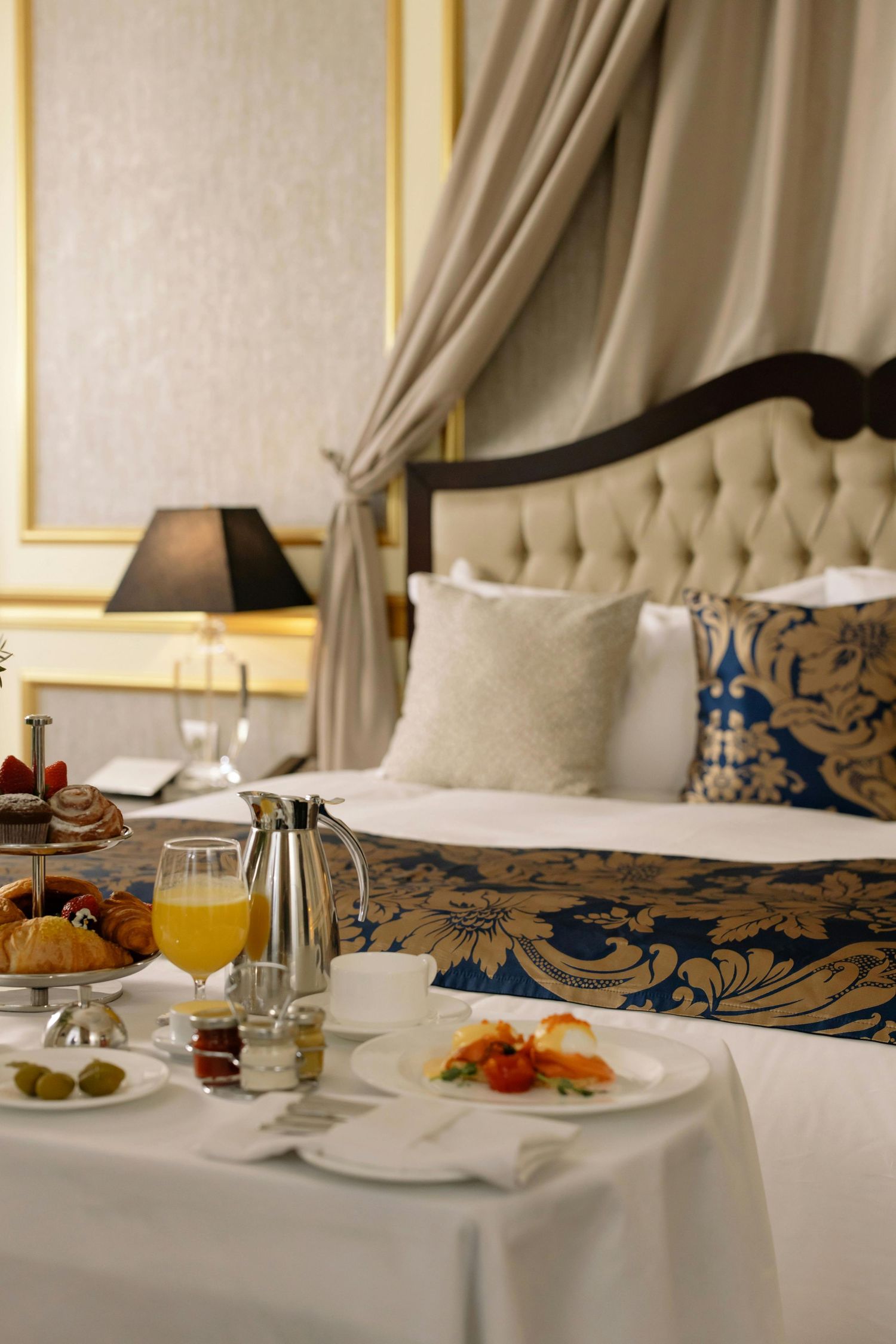 Luxury hotel room service breakfast with orange juice and pastries on white tablecloth next to elegant tufted headboard.