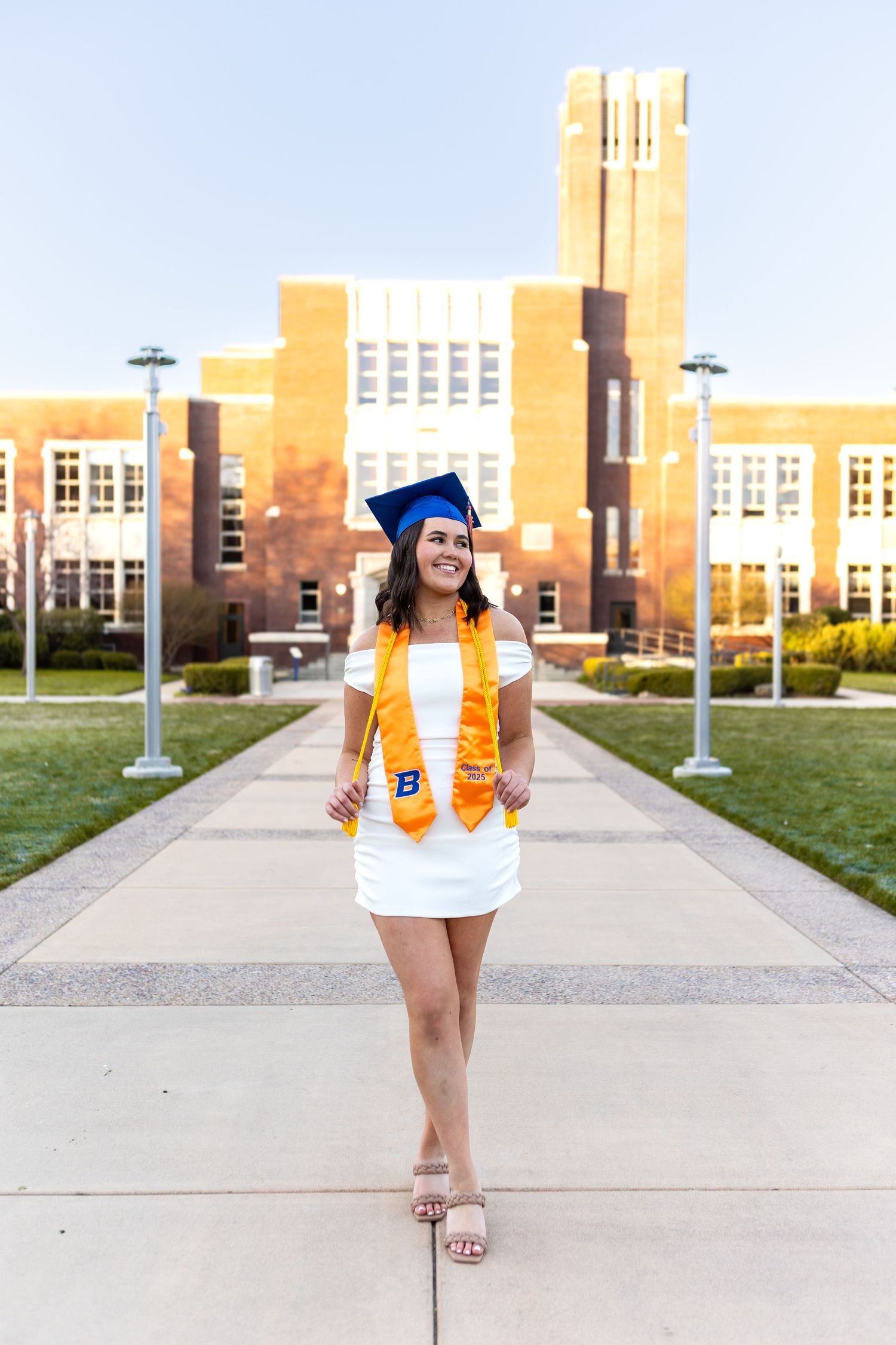 Graduate poses in cap and gown in front of a collegiate building with orange stoles and a white dress.