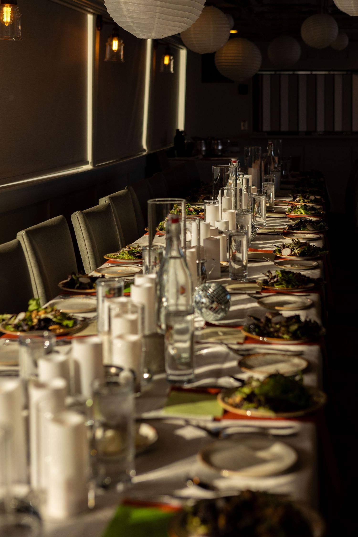 Long banquet table set for dinner with white plates, water glasses and paper lanterns hanging from dark ceiling.