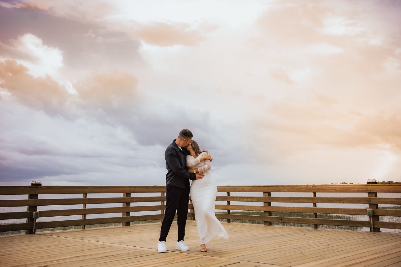 A newlywed couple embraces on a wooden deck overlooking the ocean at sunset against a dramatic pink-clouded sky.