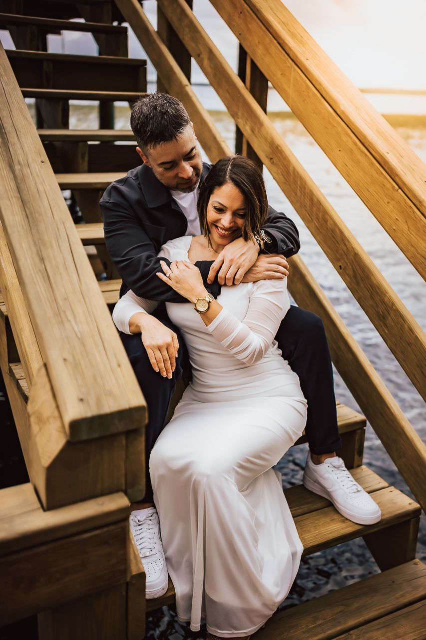 A couple in formal attire share an intimate moment while sitting on wooden outdoor stairs at sunset.