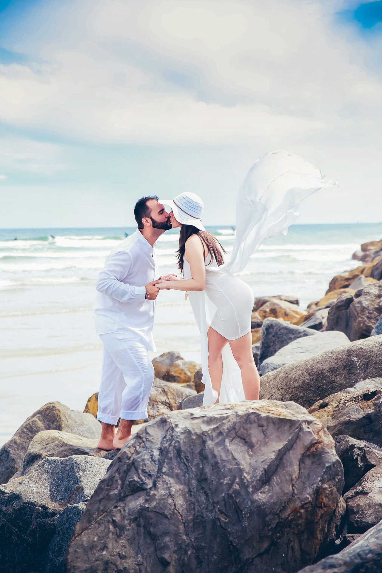 Couple in white attire share romantic moment on rocky beach as bride's veil catches ocean breeze.