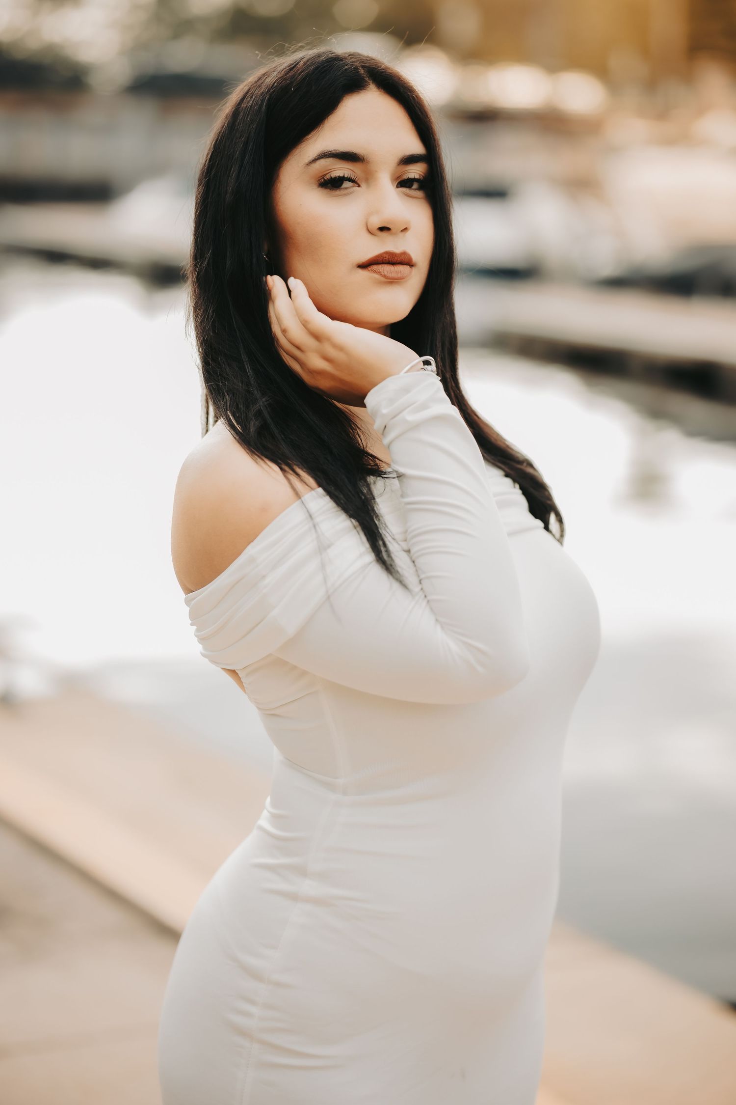 A woman in a fitted white off-shoulder dress poses outdoors in soft lighting against a blurred background.