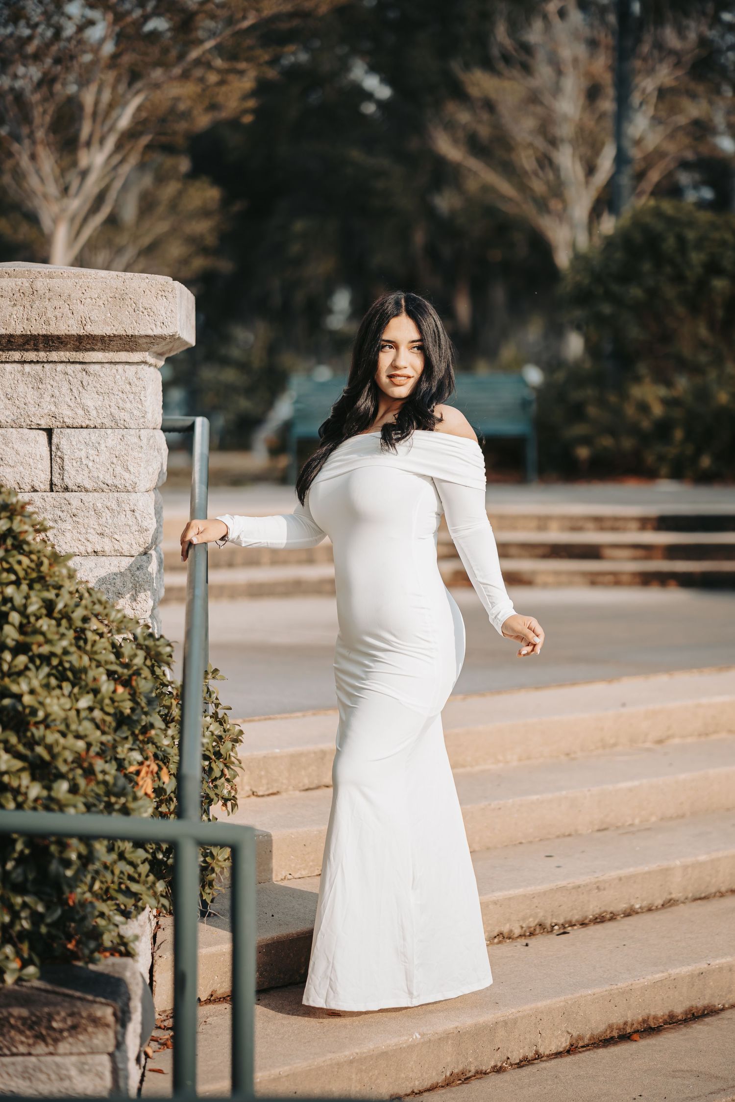 Woman in elegant off-shoulder white gown stands on stone steps in an outdoor garden setting at sunset.