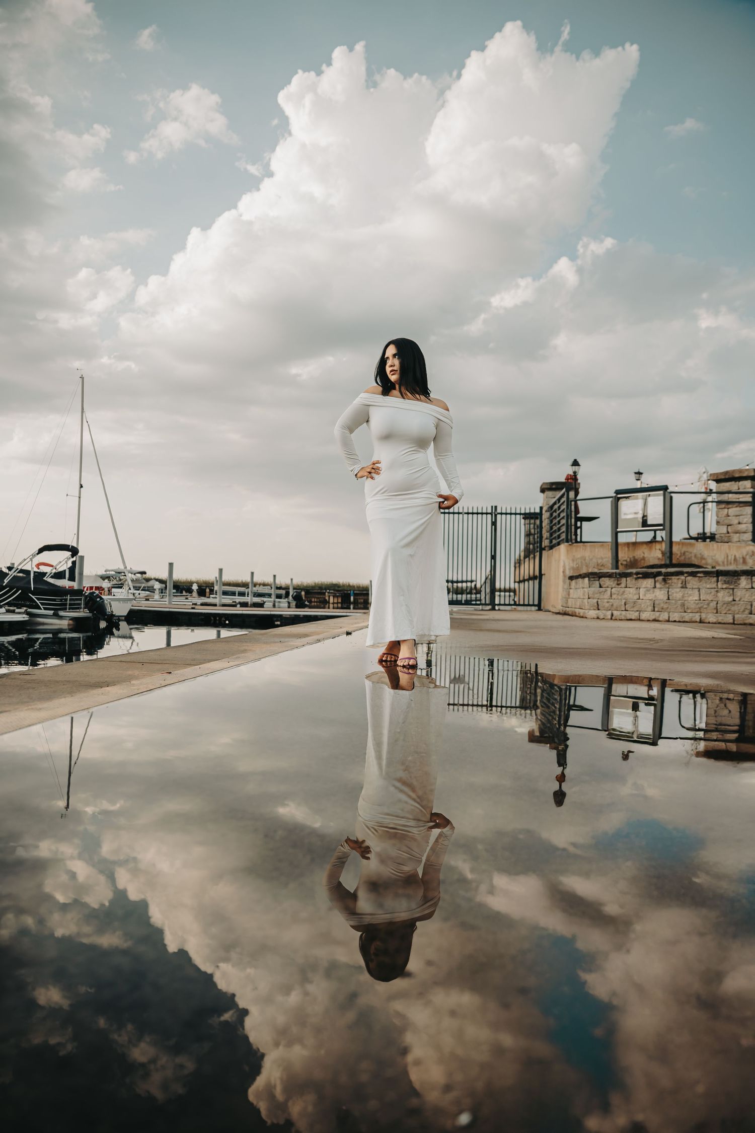 Artistic reflection photograph of woman in white dress standing by marina with dramatic clouds and water mirroring the scene.