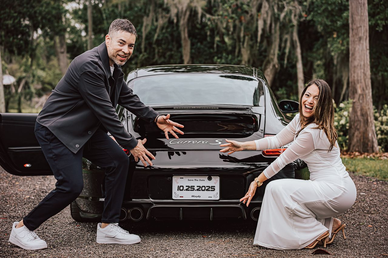 A couple wearing white outfits pose playfully in front of a black car on a dirt path surrounded by trees.