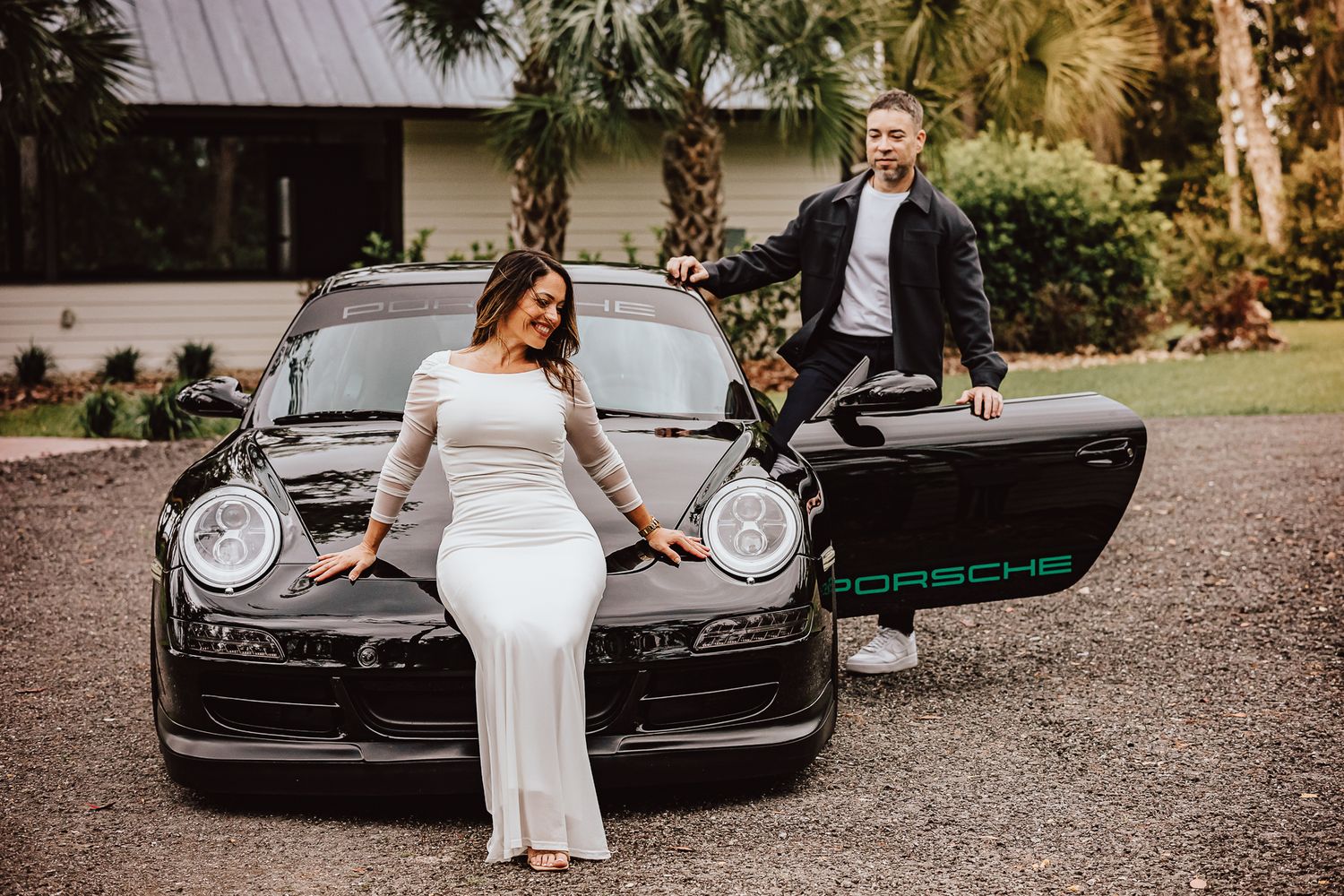 A sleek black Porsche serves as a luxury prop for a wedding photo shoot on a gravel driveway.