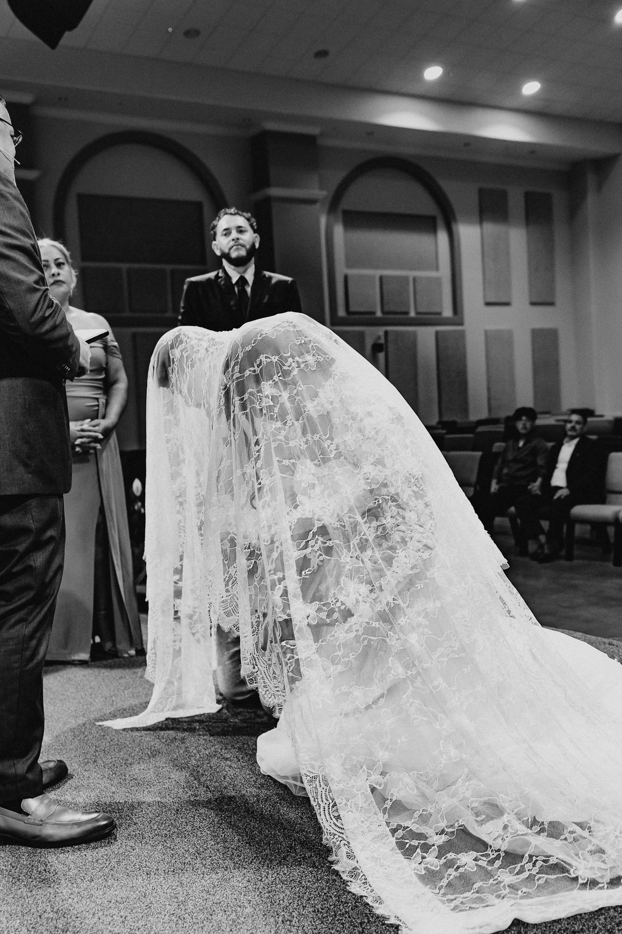 Black and white photo showing intricate lace wedding veil spread out on church floor during ceremony.