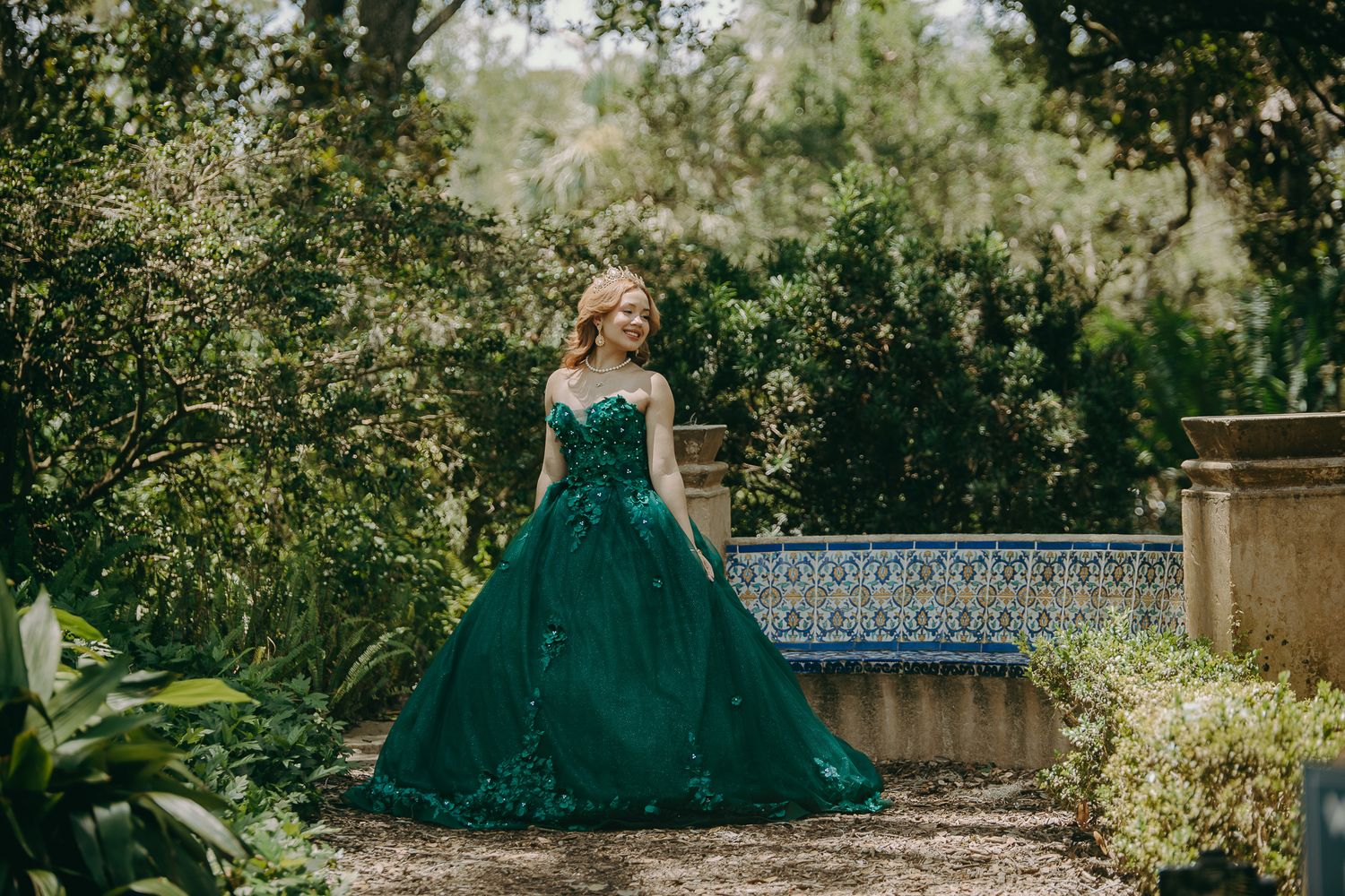 A person in an emerald green ball gown stands in a lush garden setting with decorative tiles and greenery.