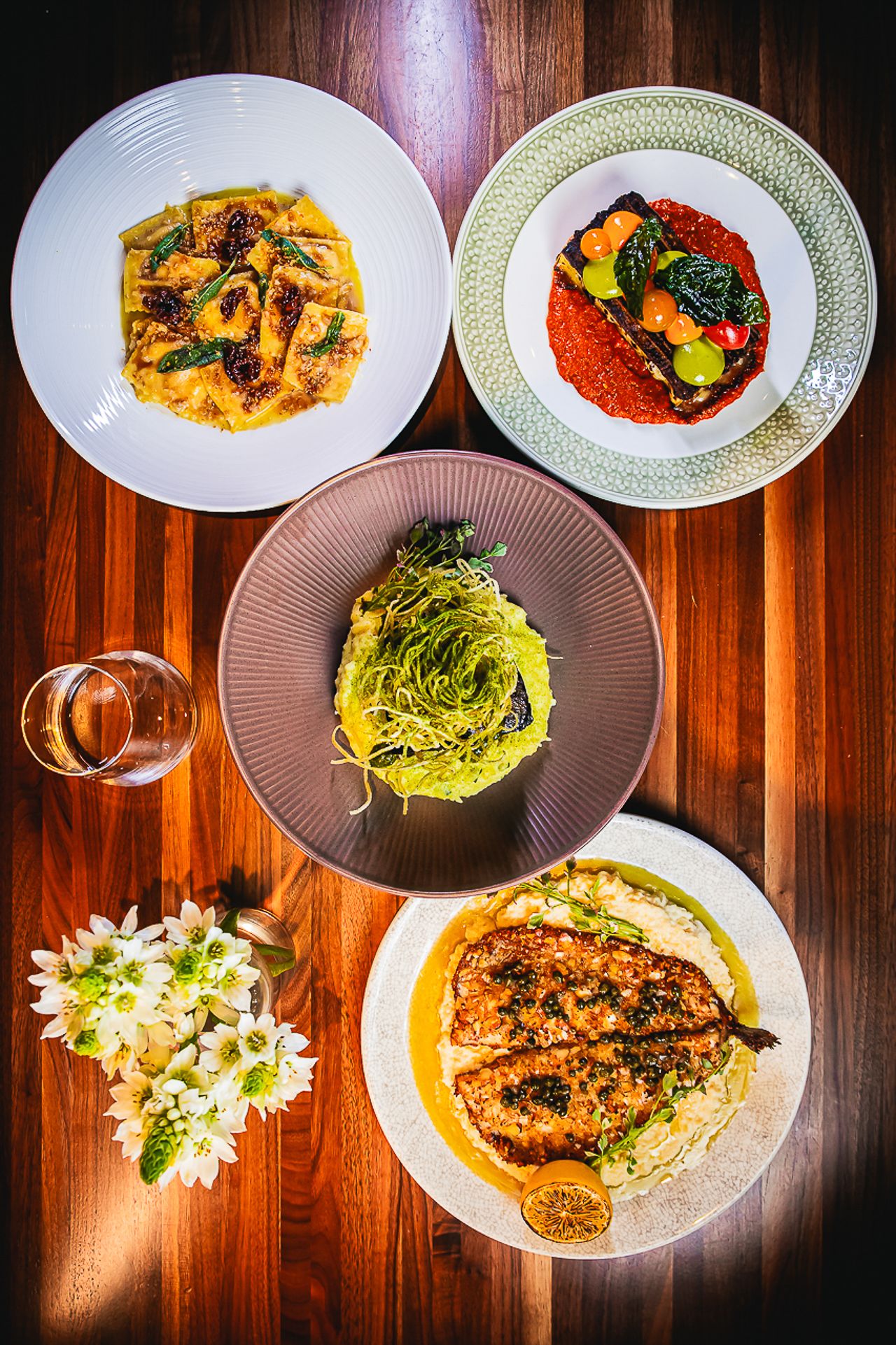 Elegant plating of four gourmet dishes including pasta, meat, and vegetables on a wooden table with white flowers.