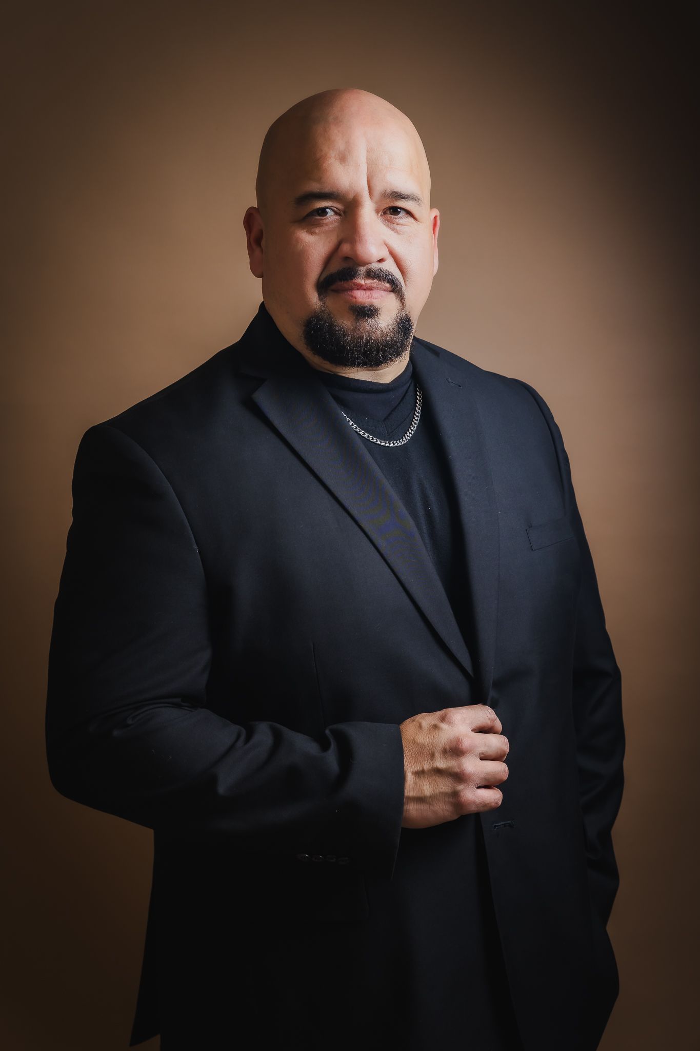 Professional studio portrait of a person in a black blazer and turtleneck against a brown background.