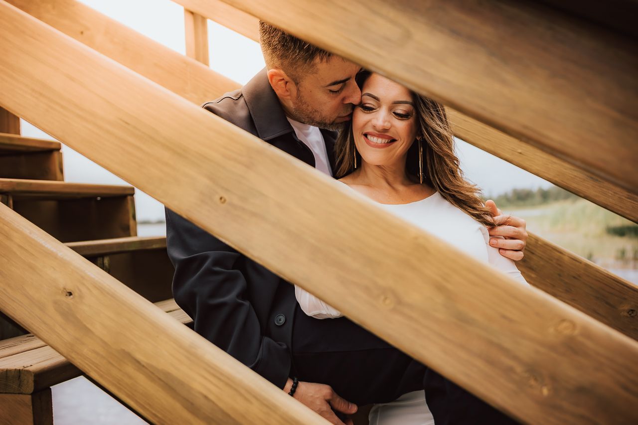 A loving couple in formalwear share an intimate moment between wooden stair railings in warm lighting during sunset