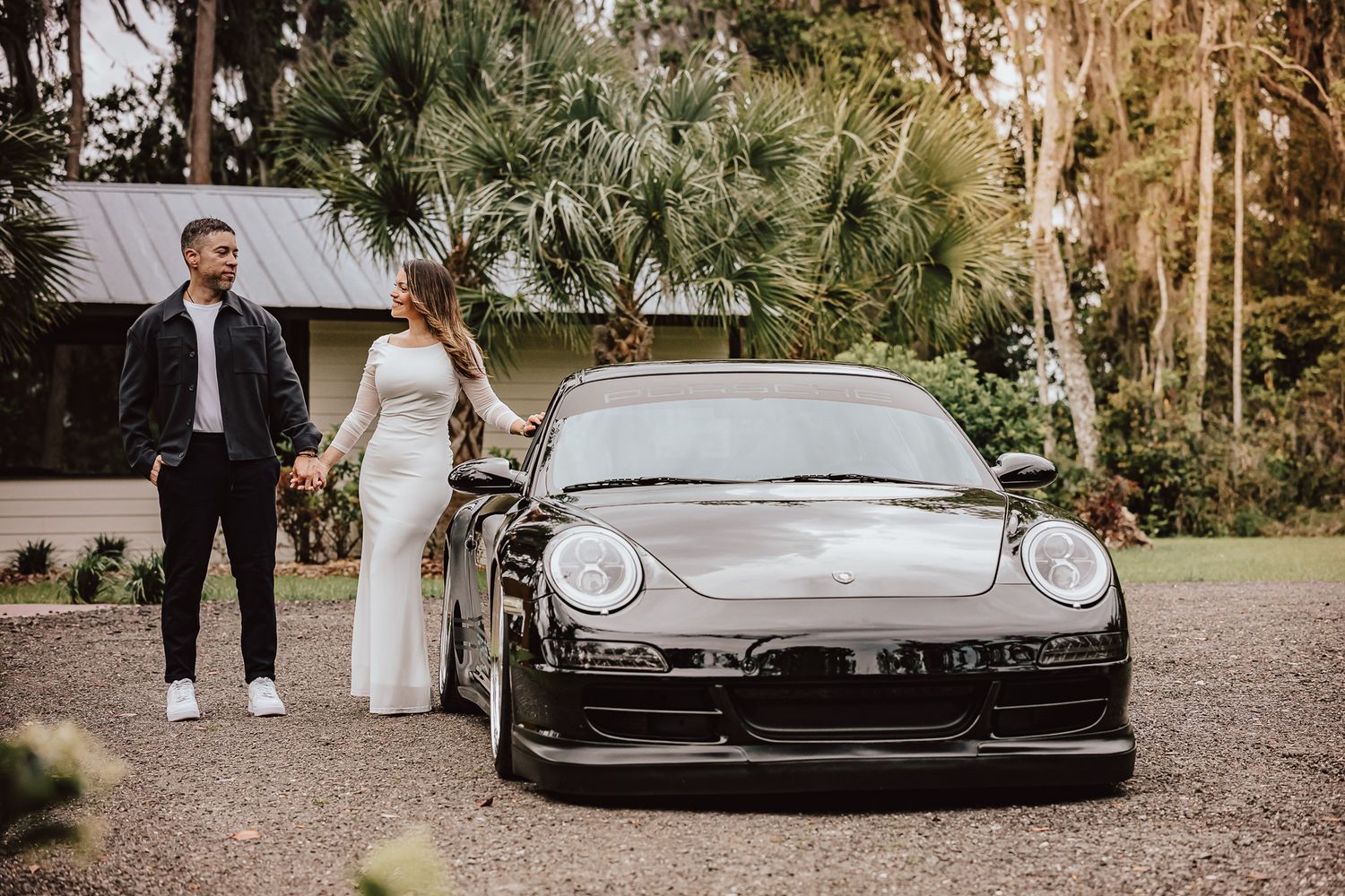 A couple stands beside a sleek black Porsche sports car on a gravel driveway surrounded by palm trees.