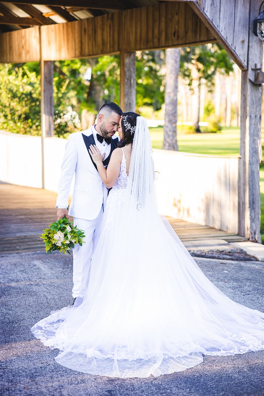 A couple in white wedding attire share an intimate moment under a wooden covered bridge with trailing veil and floral bouquet.