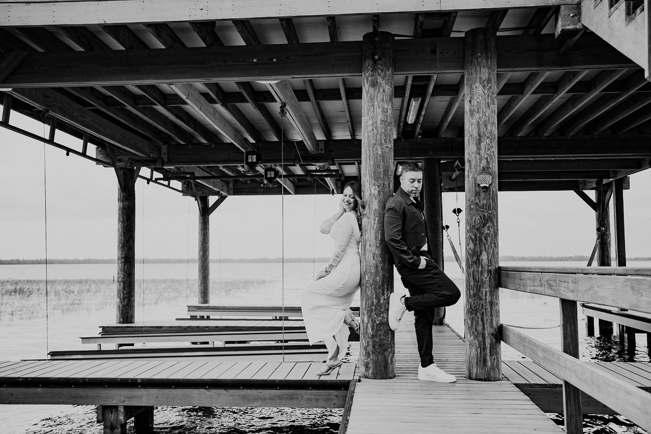 A person stands at a wooden pier overlooking a lake under a rustic covered structure in black and white.