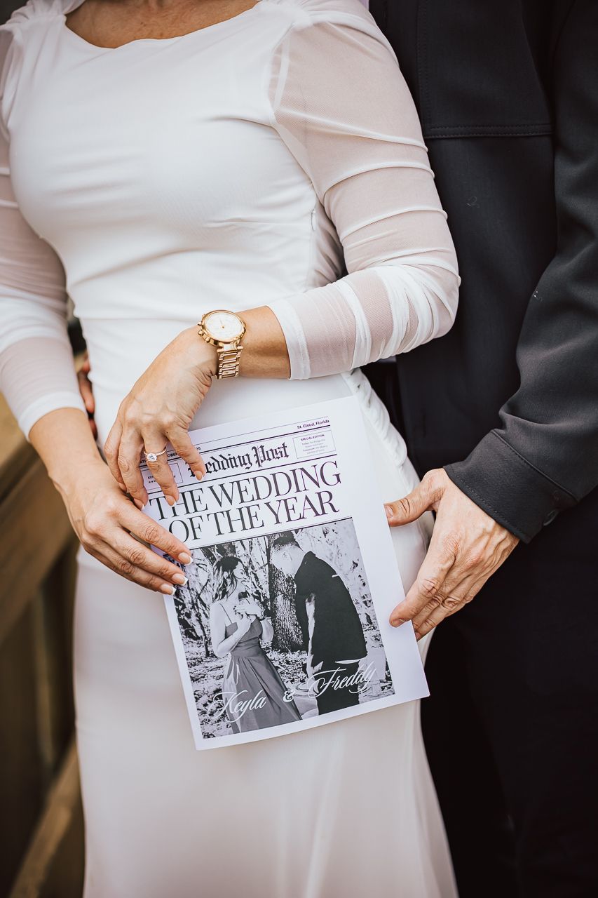 Person in white dress holding a newspaper headline titled 'The Wedding of the Year' while standing next to someone in dark attire.