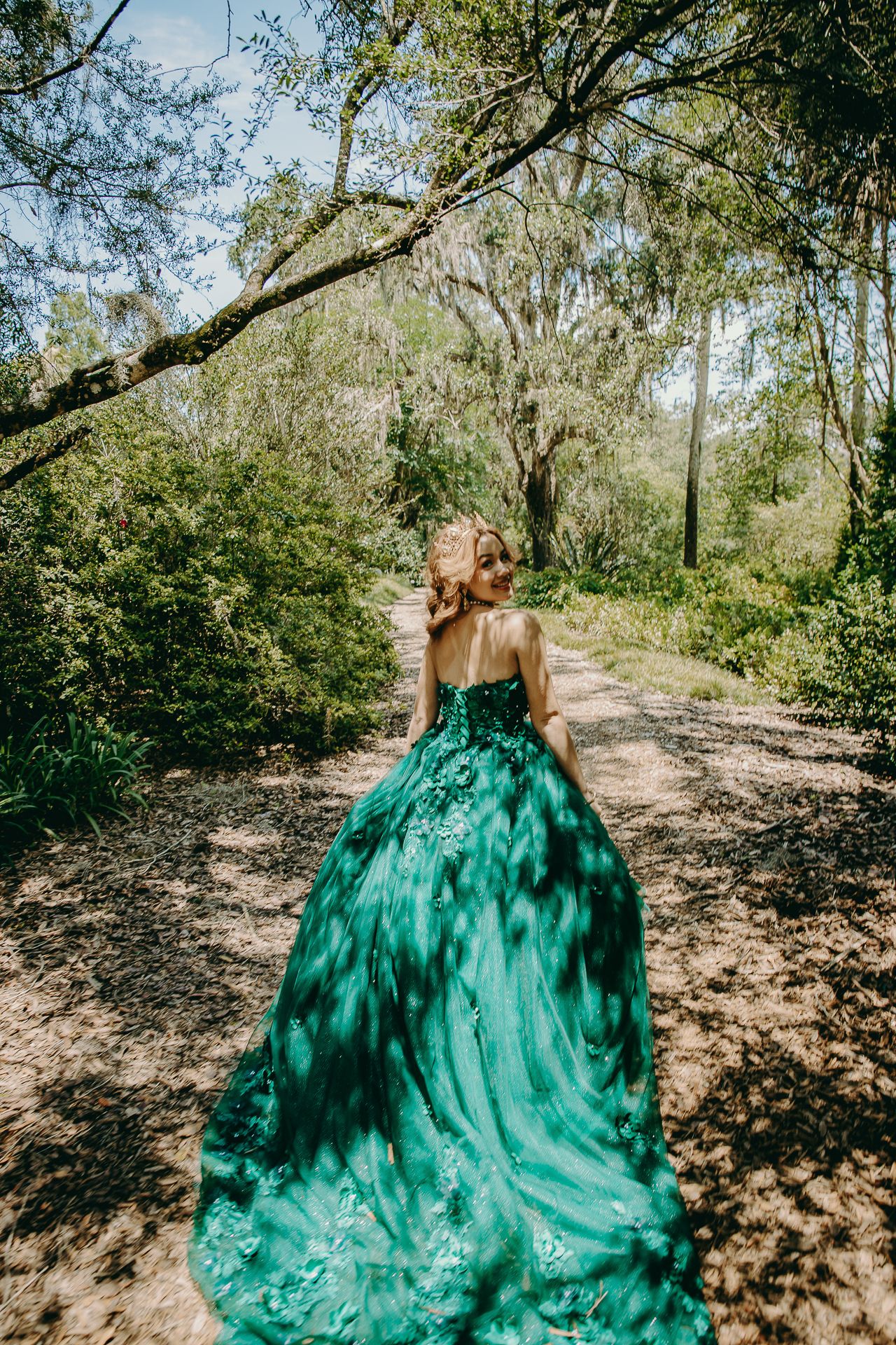 A woman in a flowing emerald green ballgown walks along a sunlit wooded path with Spanish moss in the trees.