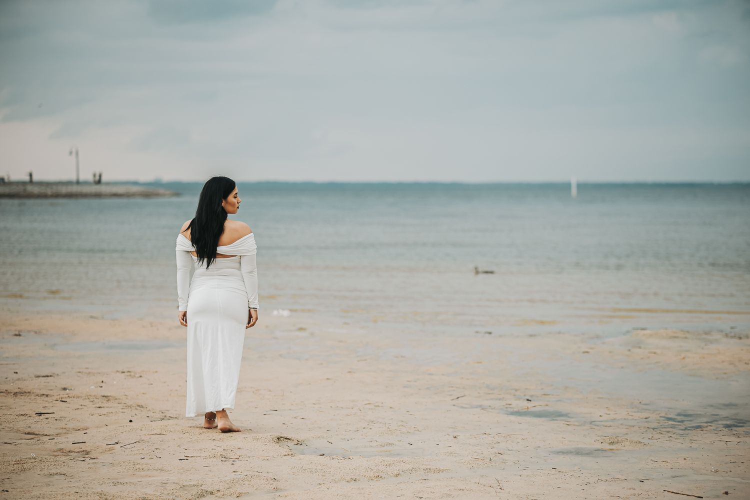 A person in a flowing white dress stands alone on a peaceful beach looking out towards the calm ocean and horizon.