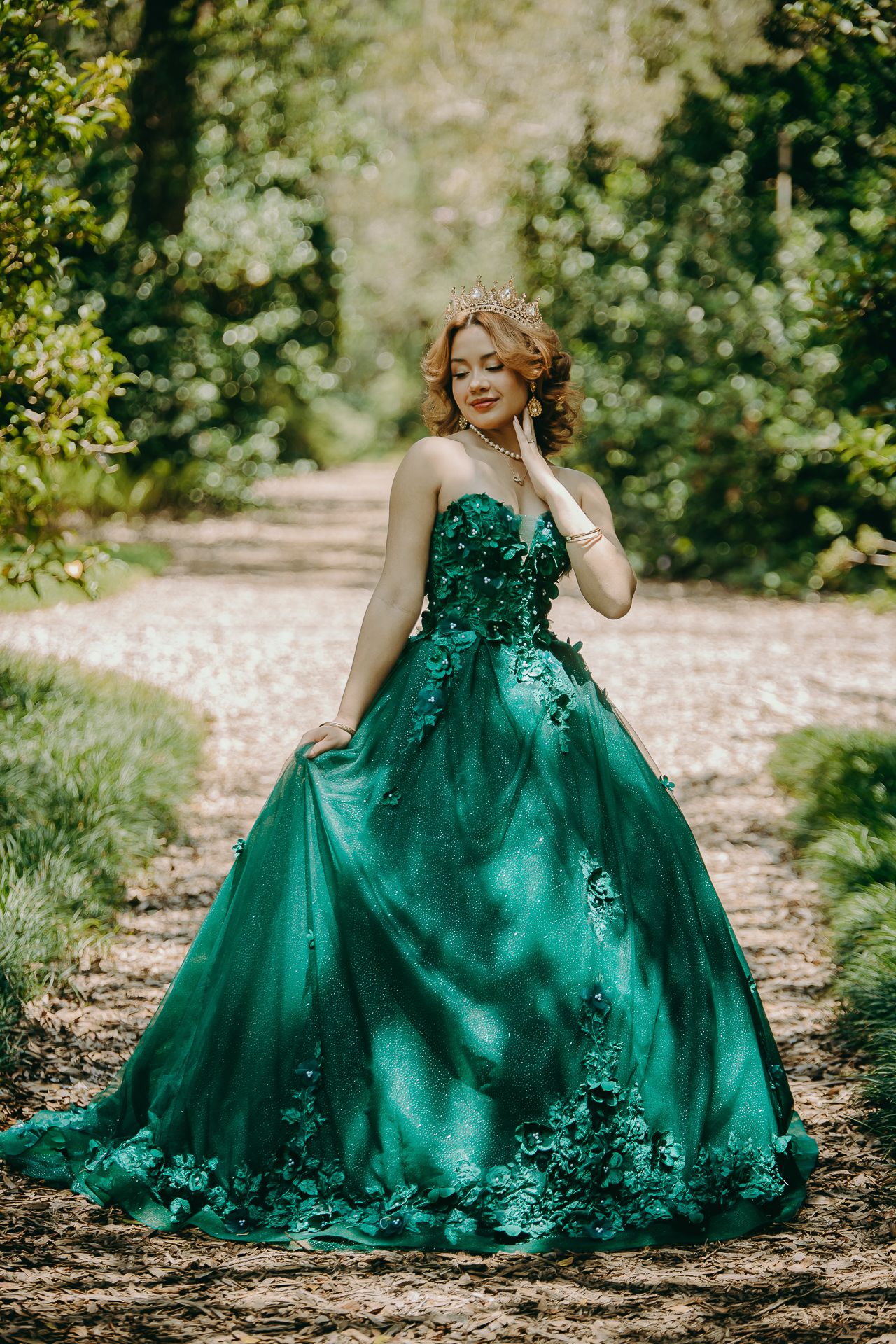 A stunning emerald green ball gown with floral appliques sparkles on a sunlit forest path.