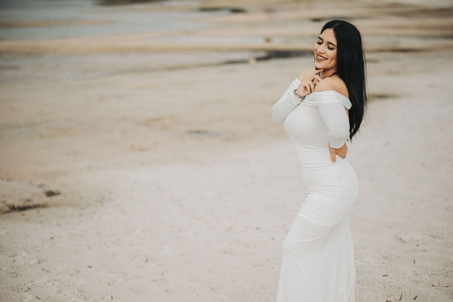 An expecting mother in a flowing white dress poses on the beach during a maternity photoshoot.