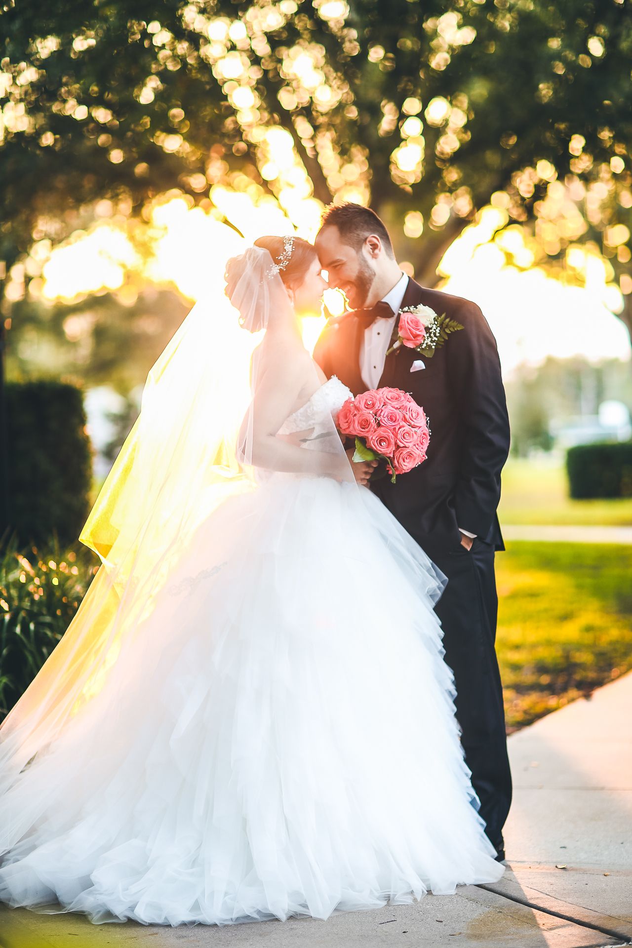 Bride and groom share intimate moment in golden sunlight with flowing veil and pink bouquet.