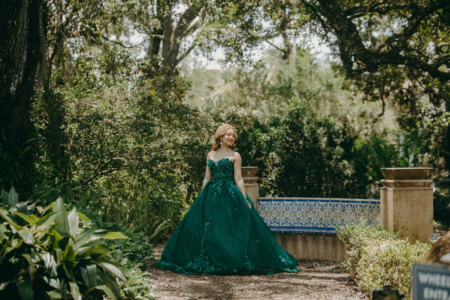 Elegant emerald green ballgown in a lush garden setting with ornate stone bench and tropical foliage.